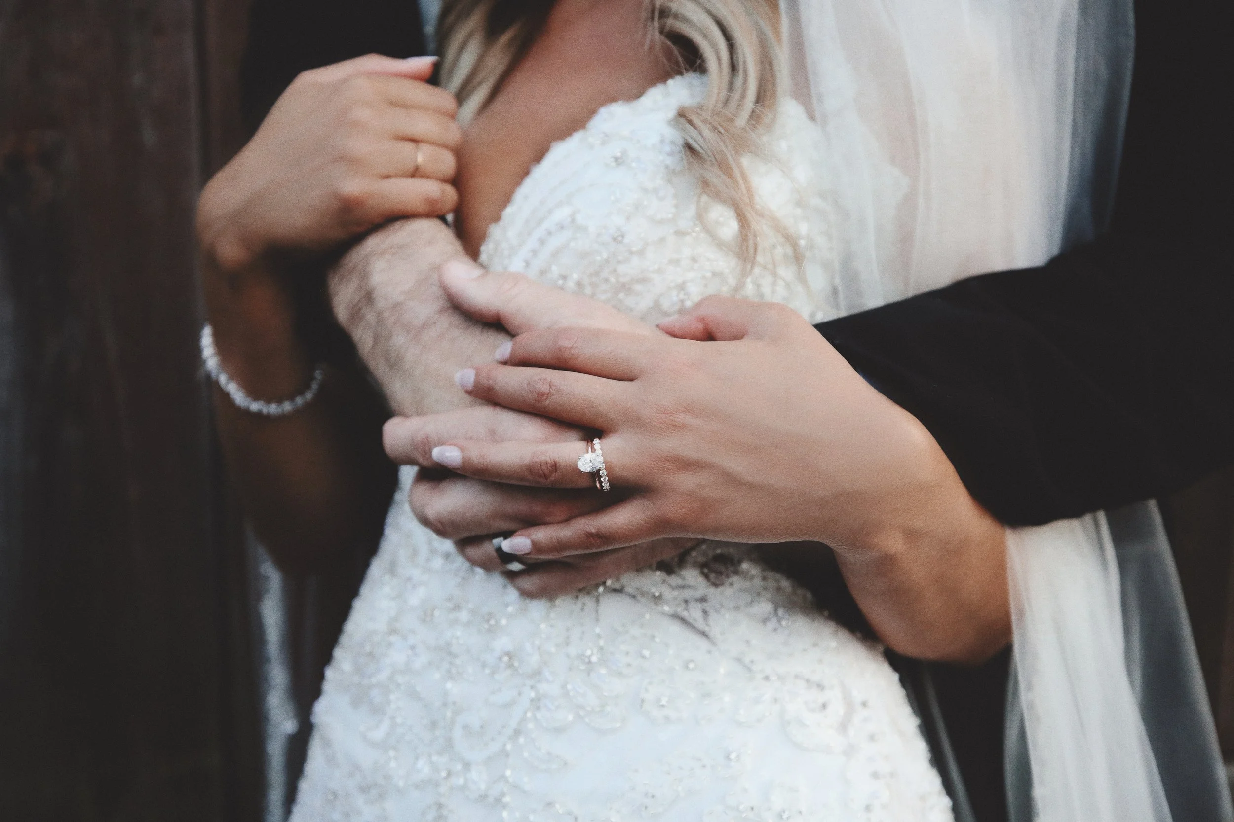 Close-up of a bride and groom embracing, with the groom's hands holding the bride's arm, displaying wedding rings.