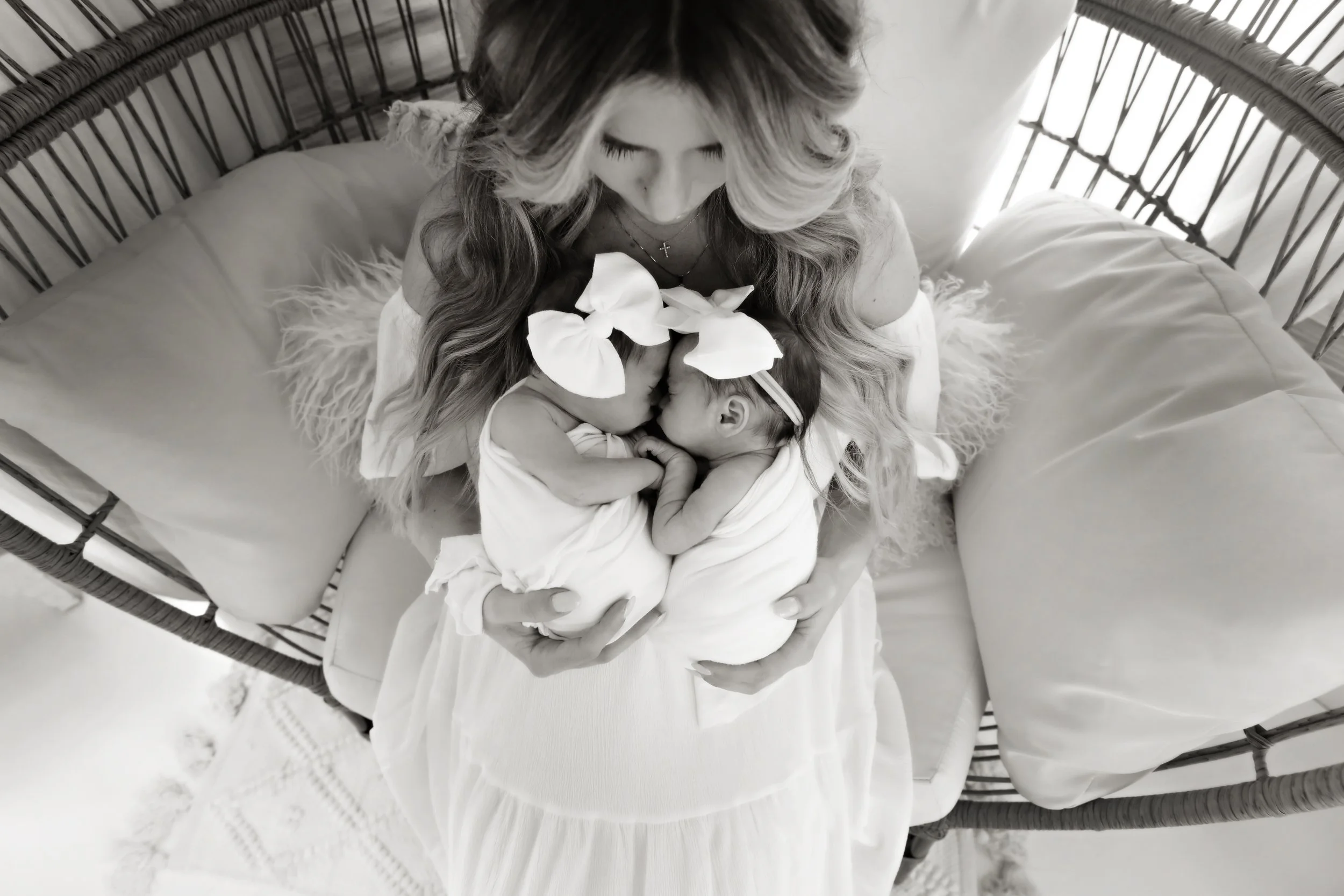 A woman with long, wavy hair and a cross necklace sitting on a cushioned chair, holding two swaddled newborn babies who are touching noses and wearing matching headbands with bows.