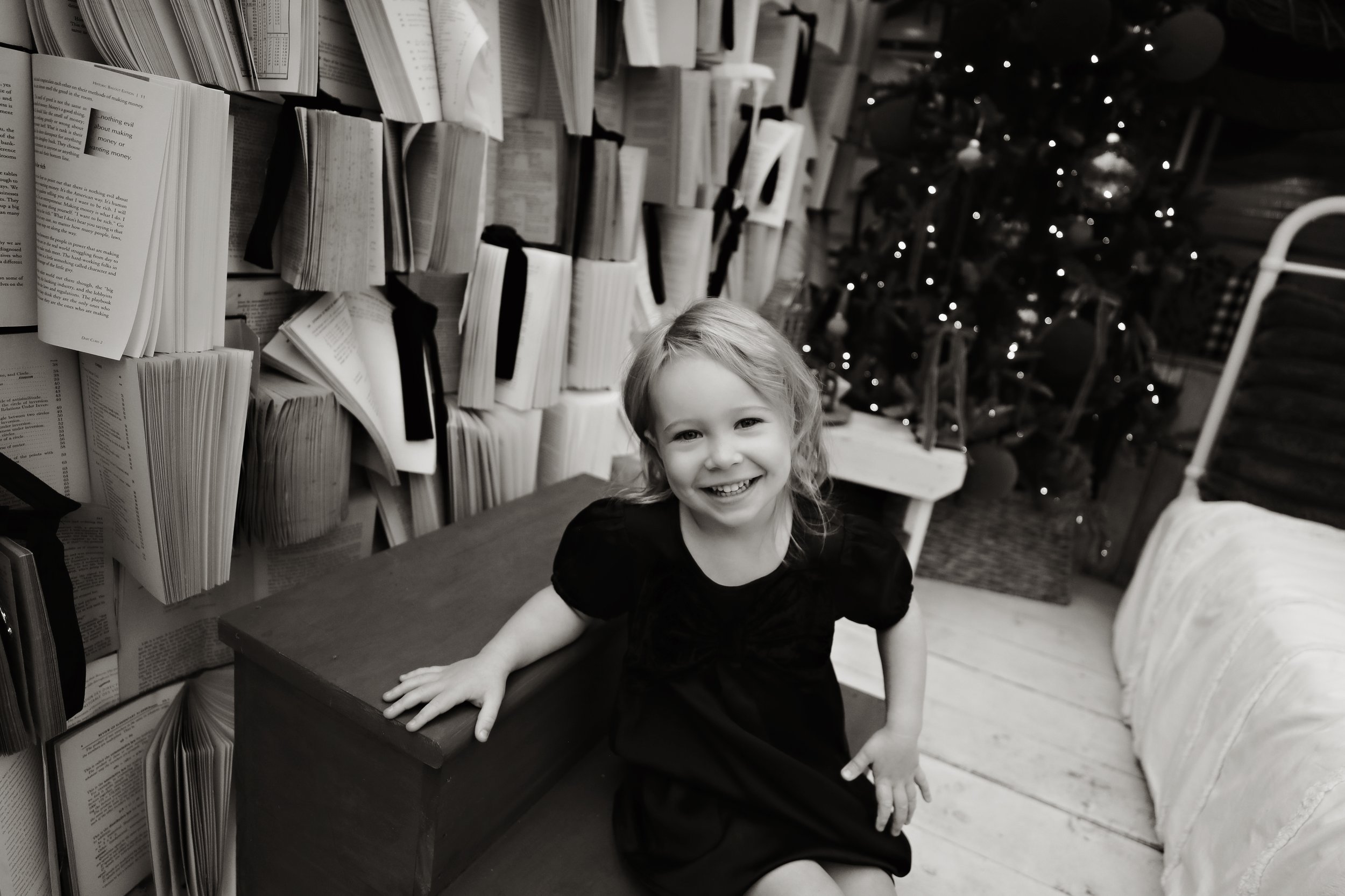 A young girl with curly hair smiling and kneeling on a bench, in front of a wall decorated with open books tied with ribbons, and a decorated Christmas tree in the background.
