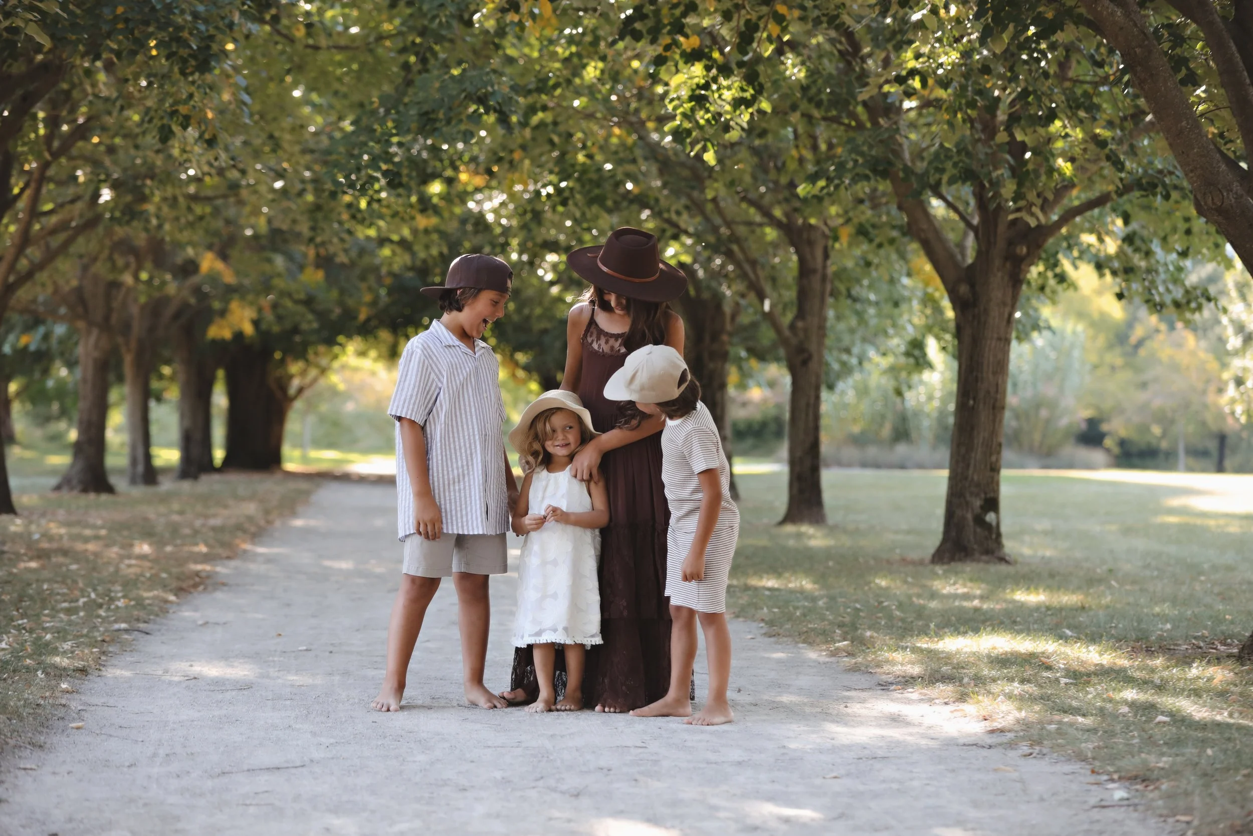 Four children and one woman standing on a dirt path in a park with trees around them, looking at each other and smiling.