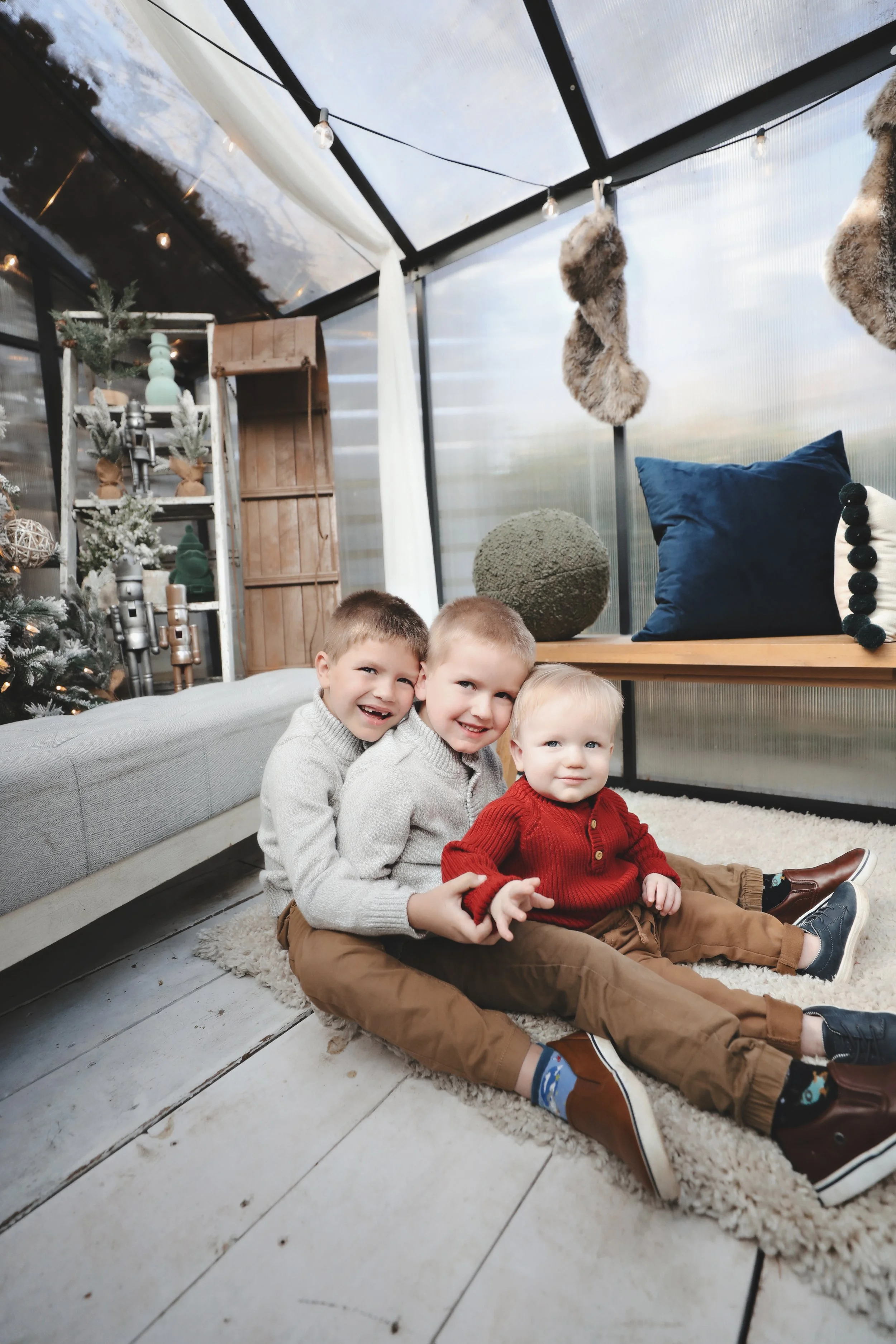 Three young boys sitting on the floor of a decorated room with Christmas trees and holiday ornaments, smiling and posing for the camera.
