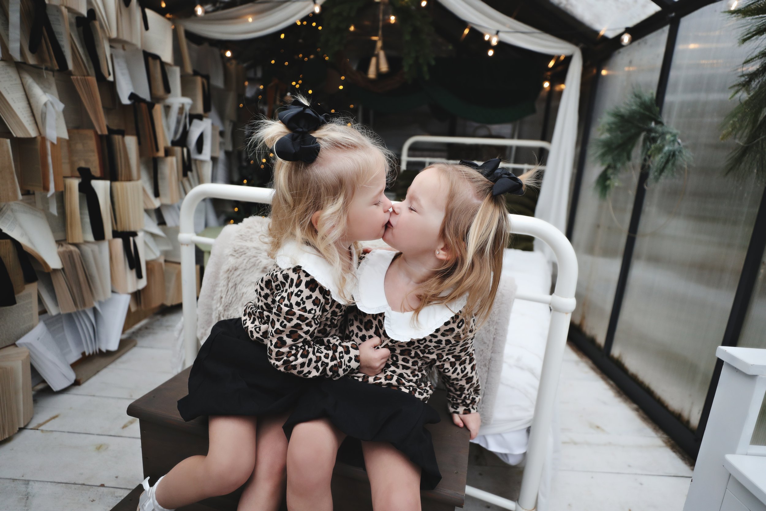Two young girls with blonde hair and black bows in their hair, dressed in leopard-print tops, are sitting on a bench and sharing a kiss in a decorated indoor space with fairy lights, books, and greenery.