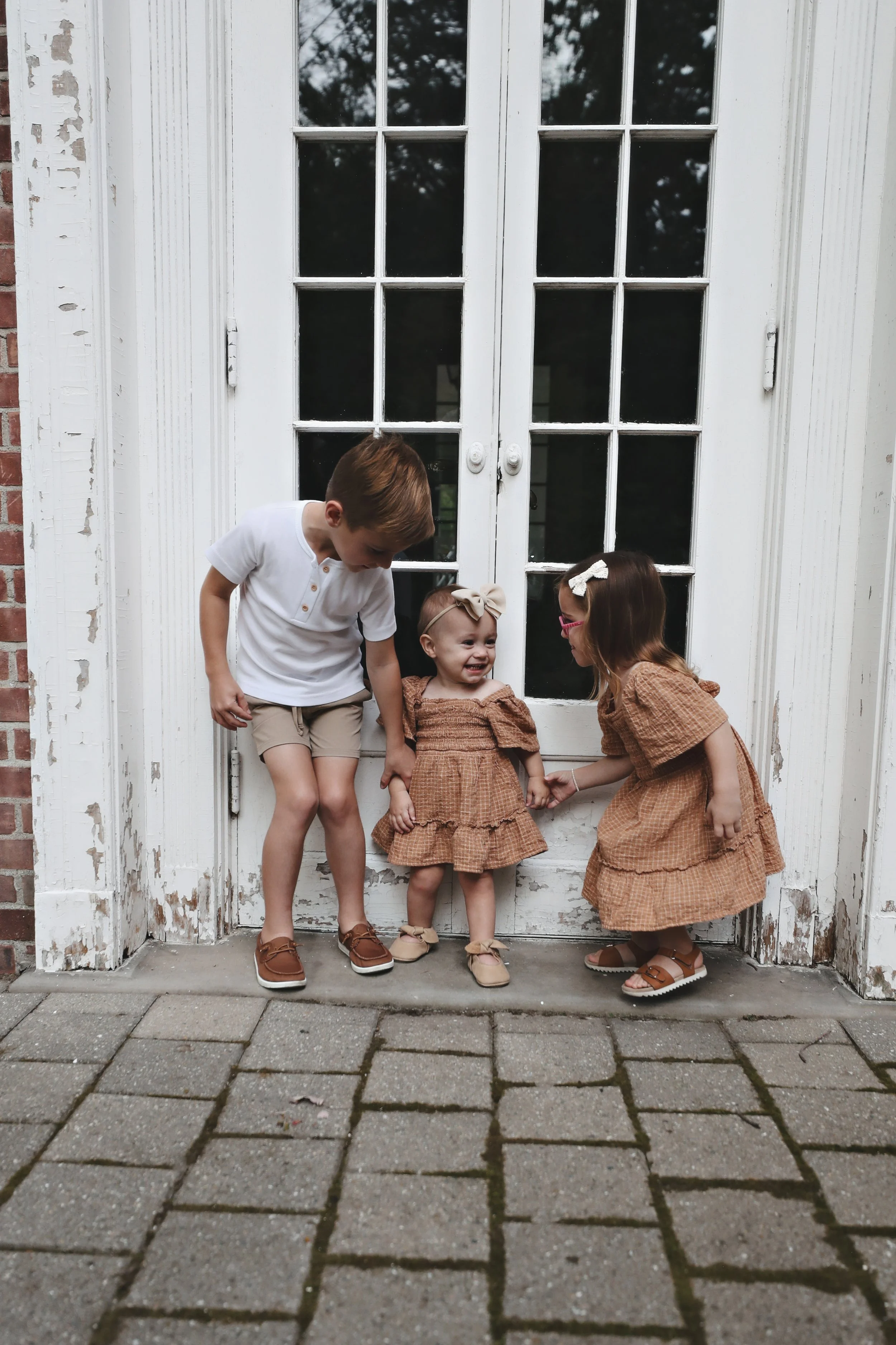 Three children, a boy and two girls, holding hands and smiling, standing on a brick sidewalk in front of a white door with glass panes and peeling paint, with trees reflected in the glass.