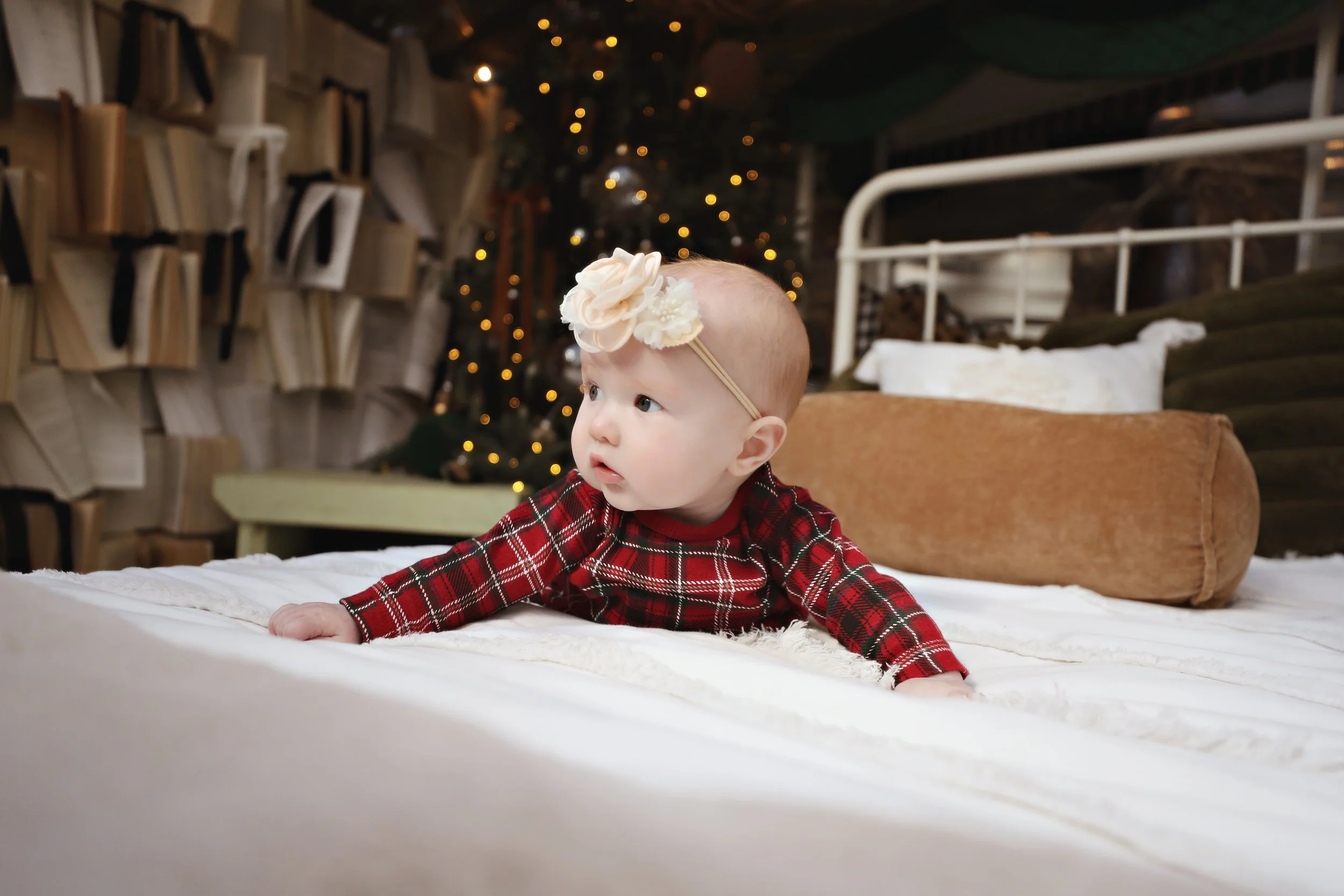 A baby girl on her hands and knees on a bed, wearing a red plaid dress and a flower headband, in a cozy room decorated for Christmas with a tree and string lights in the background.