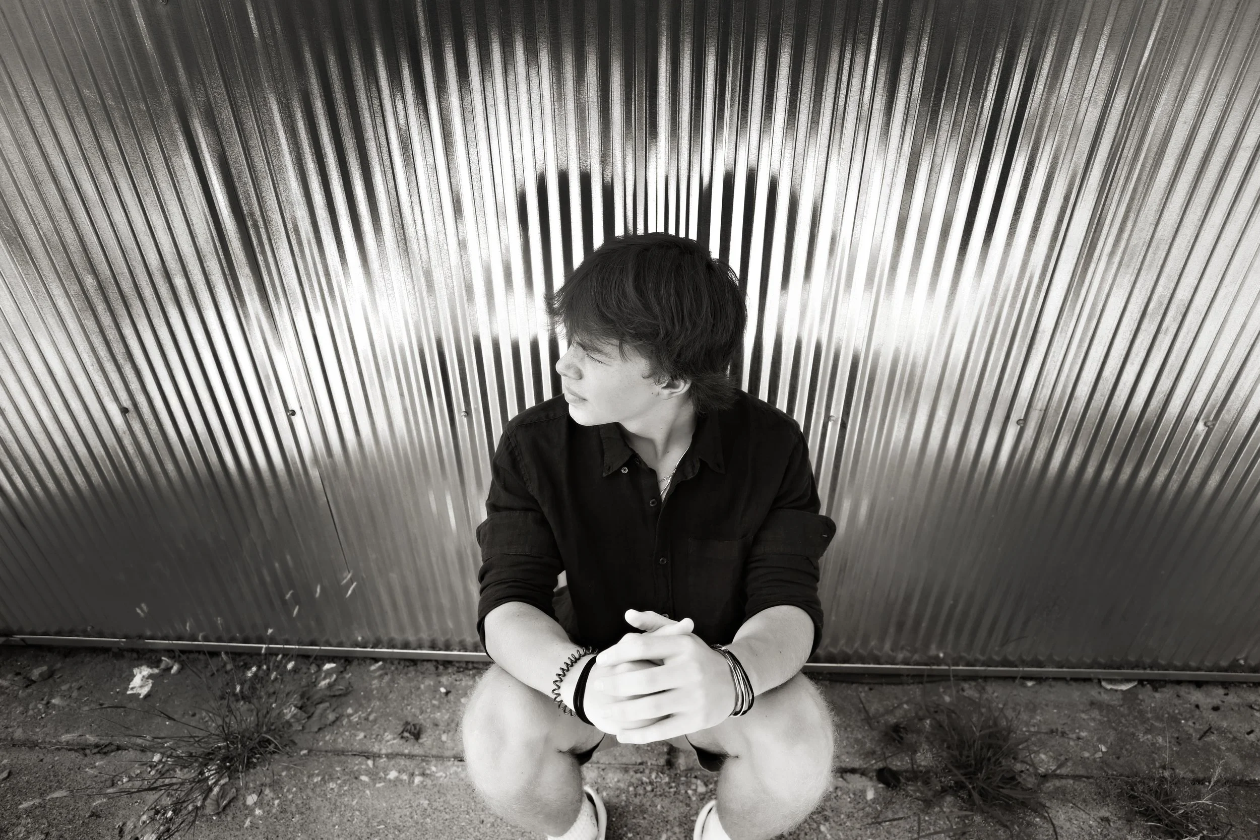 Young man with black hair sitting against a metallic, vertically ridged wall, looking to the side, wearing a black shirt and bracelets.