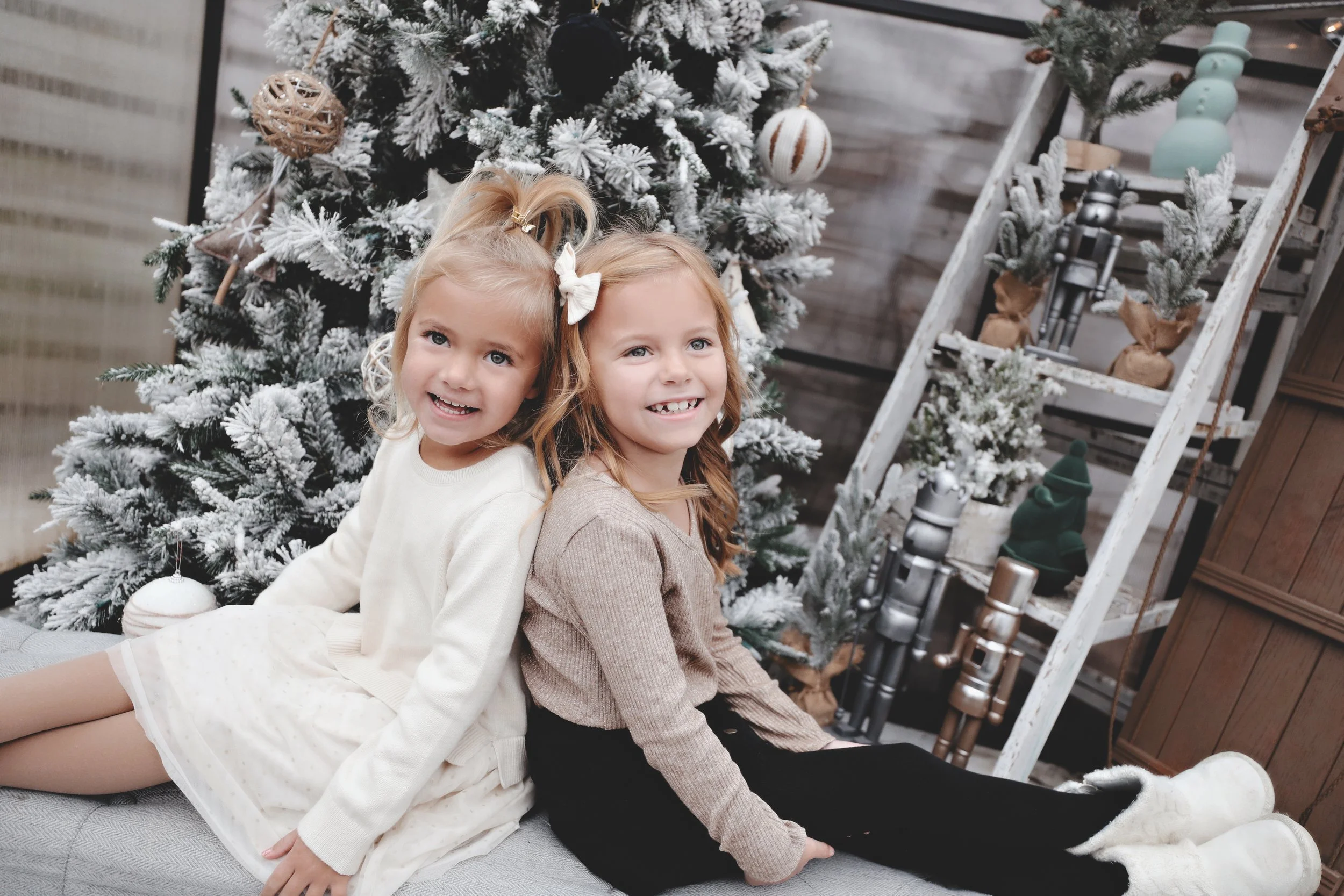 Two smiling young girls are sitting back-to-back on a gray bench, in front of a decorated snow-dusted Christmas tree, with a wooden ladder shelf with ornaments and small Christmas trees behind them.
