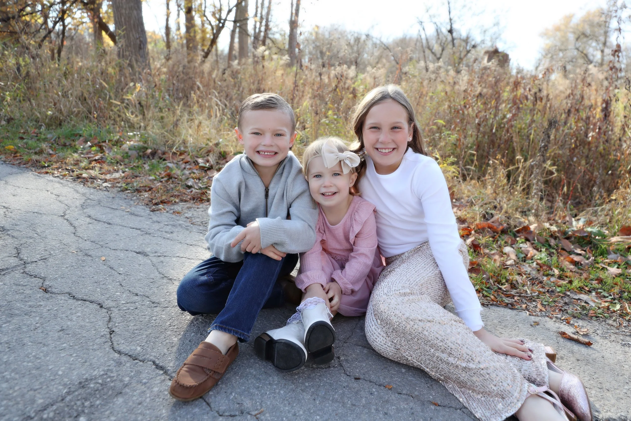 Three children sitting on the ground outdoors in a natural setting with trees and fallen leaves, smiling at the camera.