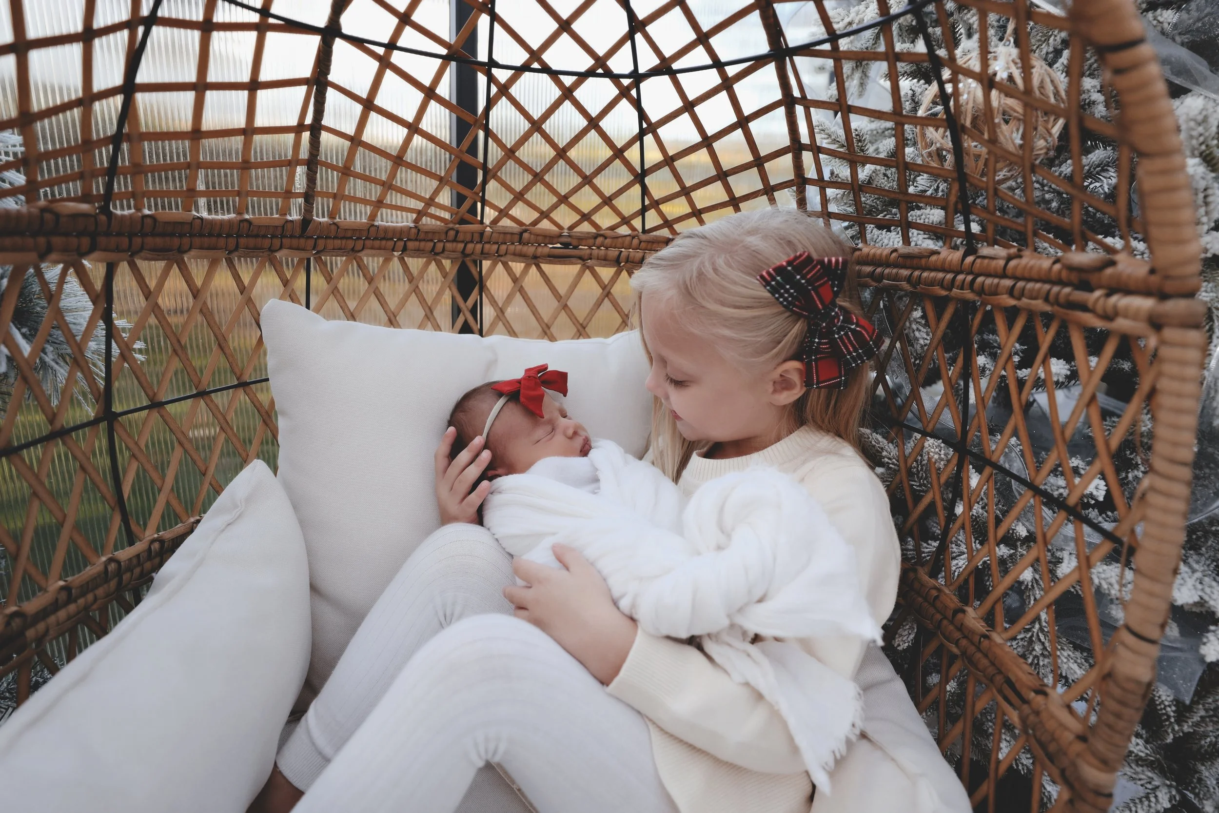 A young girl holding and looking at a sleeping baby wrapped in a blanket inside a wicker pet bed. Both are wearing matching headbands with bows.