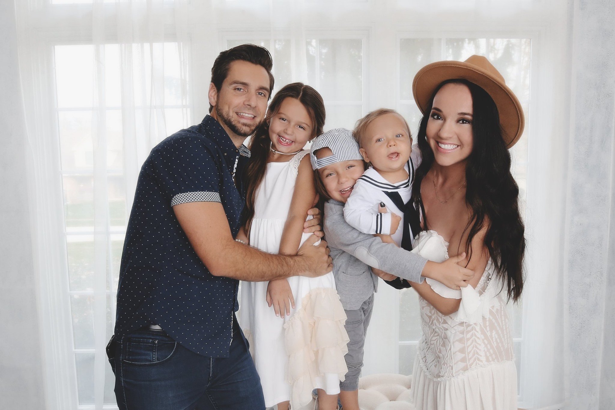 A happy family of five, including a man, woman, and three children, all smiling and standing in front of large windows with white curtains.