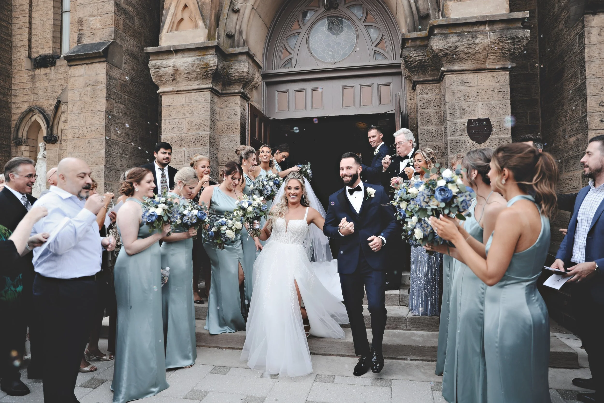 A wedding celebration with a bride and groom exiting a church surrounded by friends and family, some holding bouquets and taking photos.