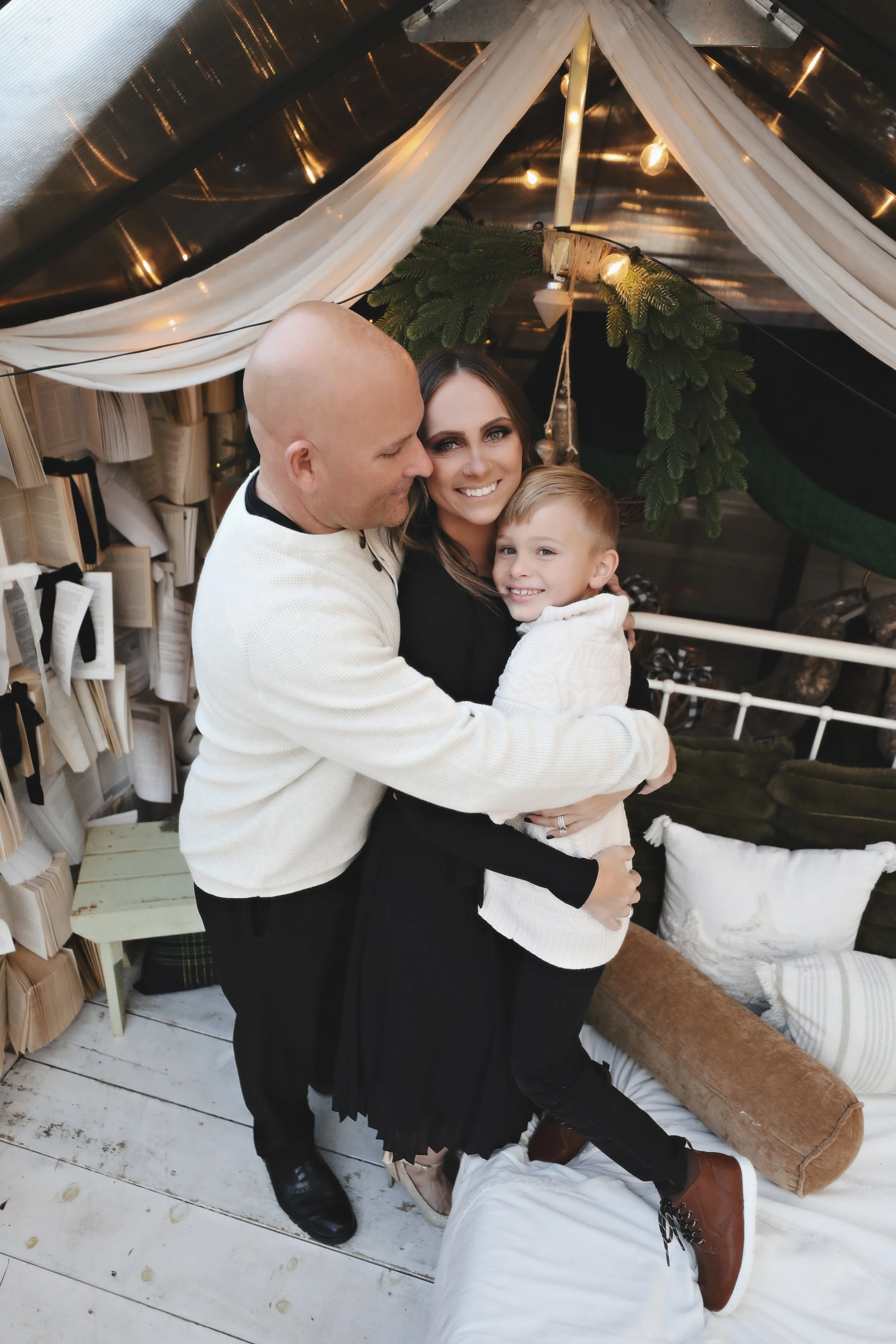 A family of three hugging inside a cozy, decorated space with books, pillows, and greenery, smiling warmly at the camera.