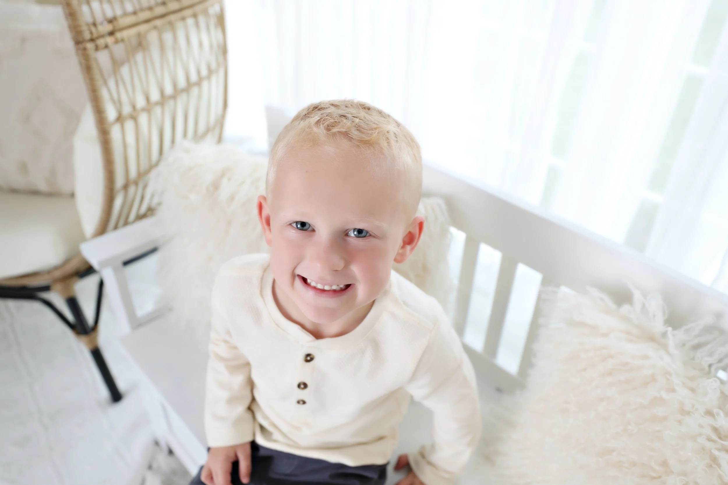 Young boy with blonde hair and blue eyes smiling while sitting on a white bench with fluffy pillows, in front of a window with sheer white curtains.