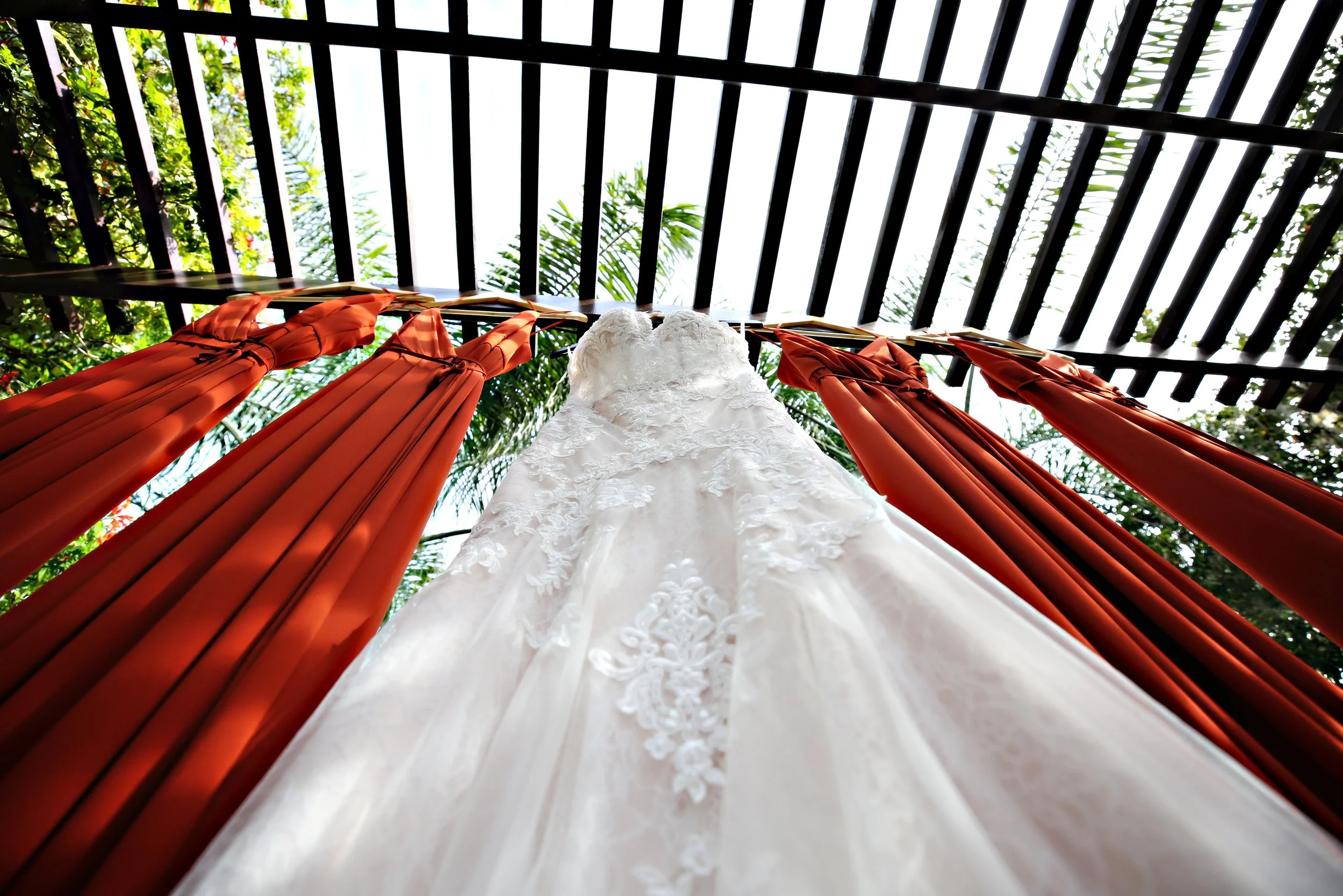 A white wedding dress hanging between orange curtains with a wooden fence and green foliage overhead in the background.
