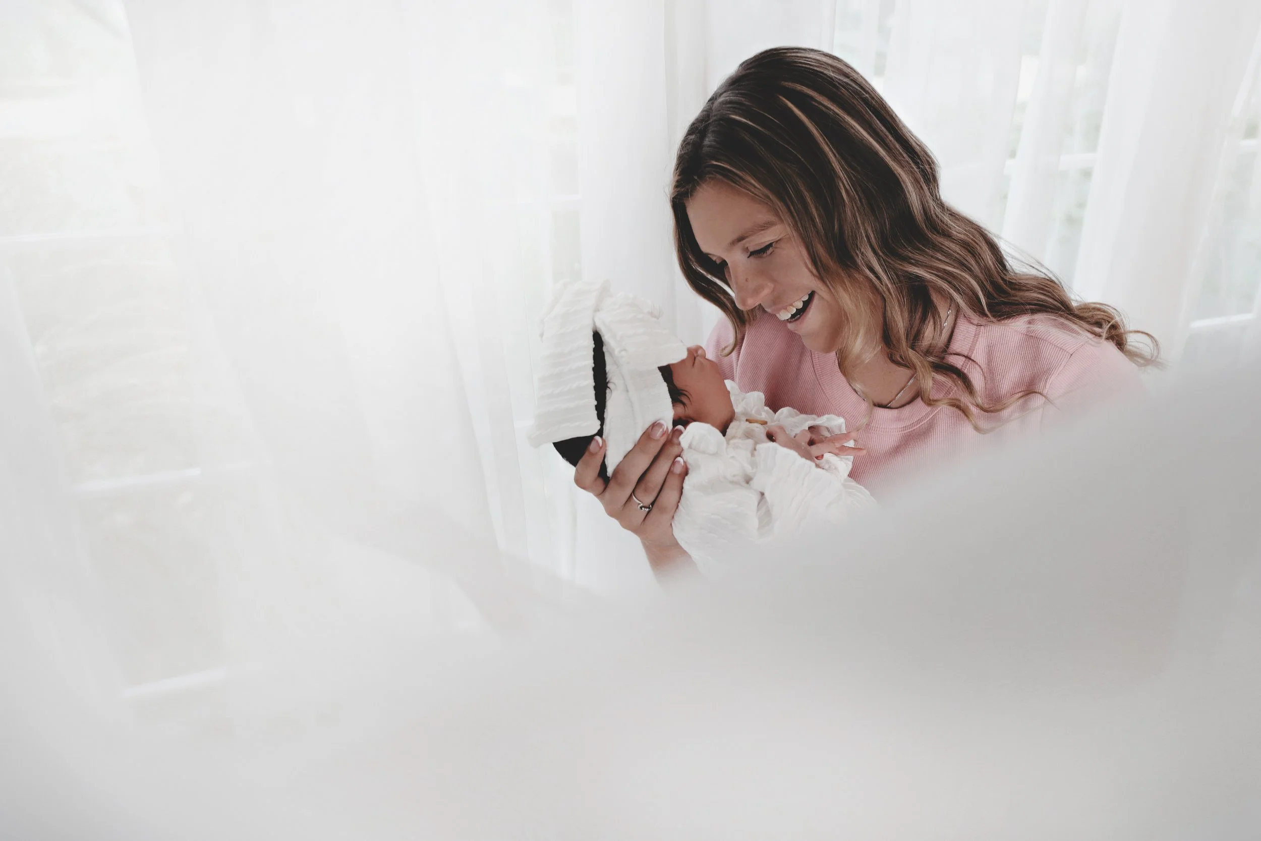 A woman holding a newborn baby with a hat, looking at the baby and smiling warmly indoors near a window with white curtains.
