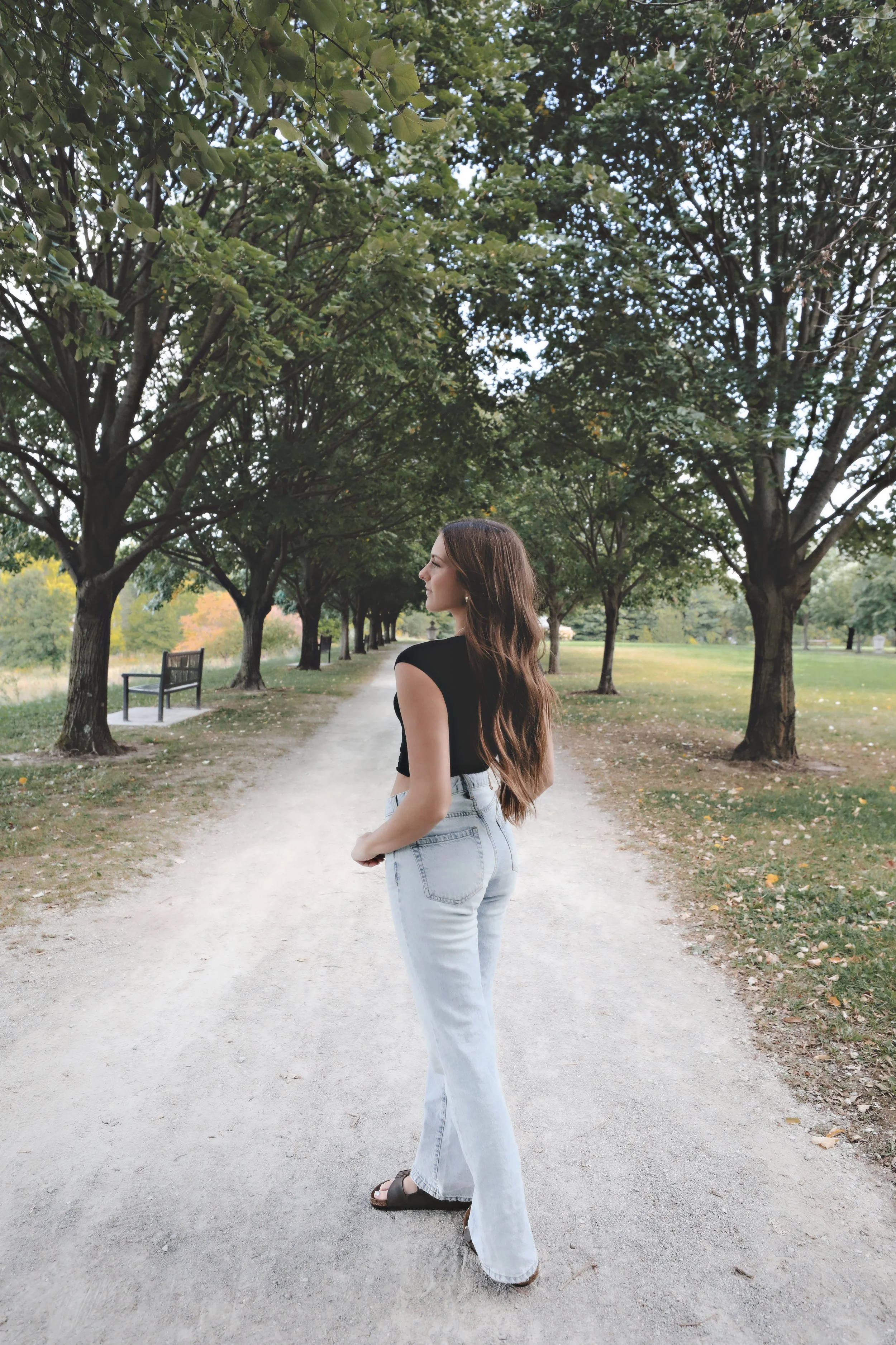 A woman with long brown hair, wearing a black sleeveless top and light-wash jeans, standing on a tree-lined gravel path in a park, looking to her left.