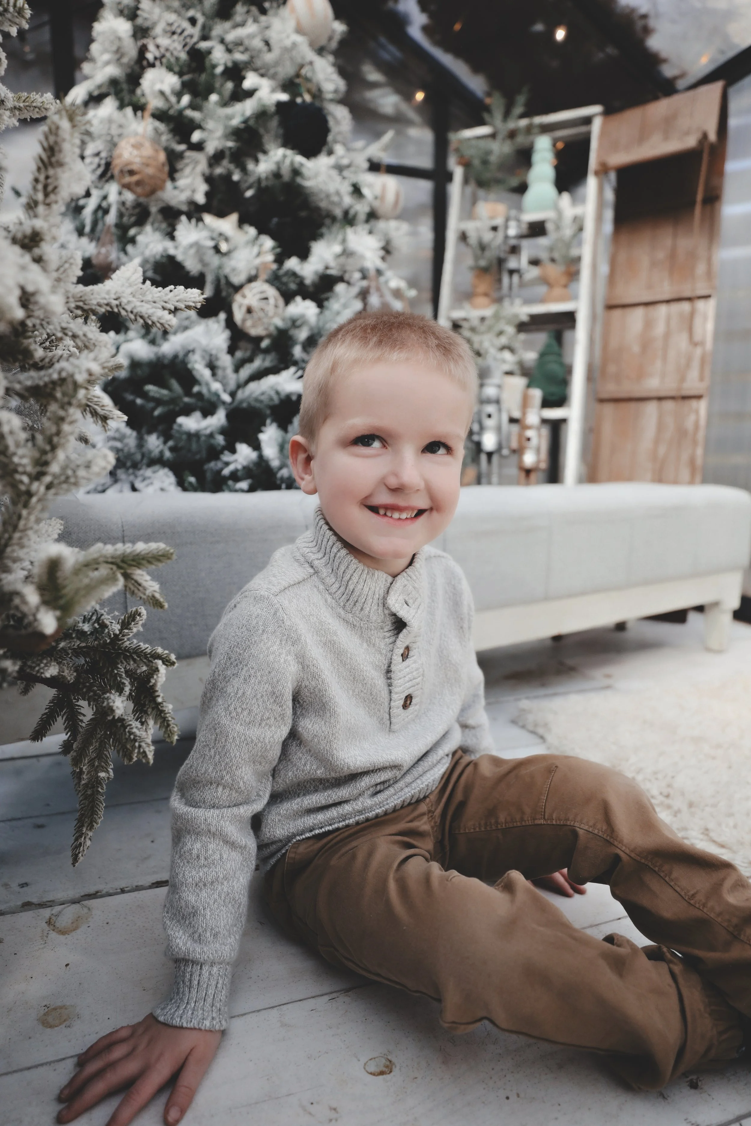 A young boy with short blond hair, wearing a gray sweater and brown pants, is sitting on a wooden floor in front of a Christmas tree decorated with ornaments and snow-like flocking, inside a cozy room with rustic decor and a white couch in the backgr