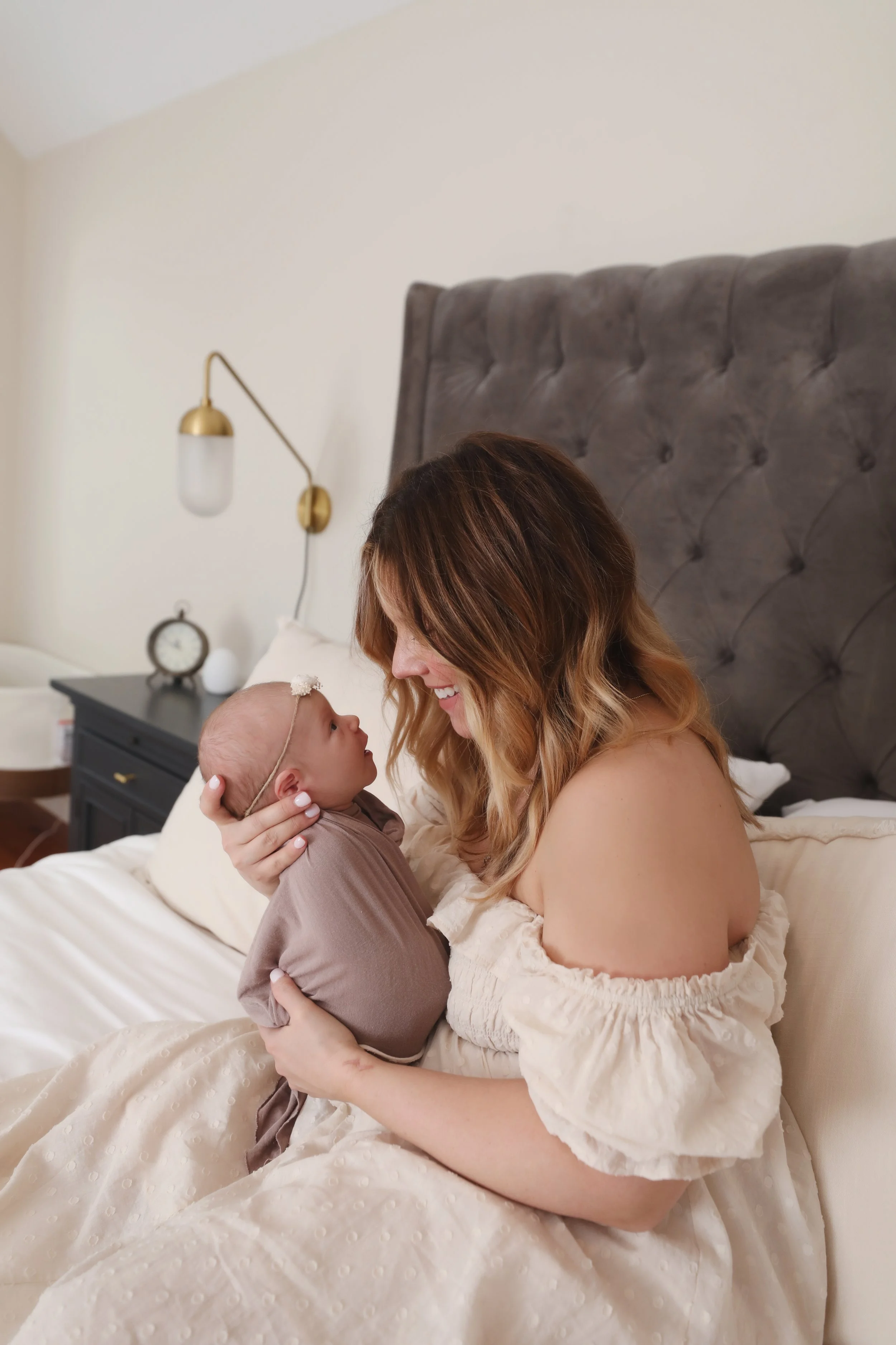 A woman with shoulder-length, wavy auburn hair holding a newborn baby in a cozy bedroom while sitting on a bed. The woman is smiling warmly at the baby, who is dressed in a brown outfit and wearing a delicate headband. The background includes a bedsi