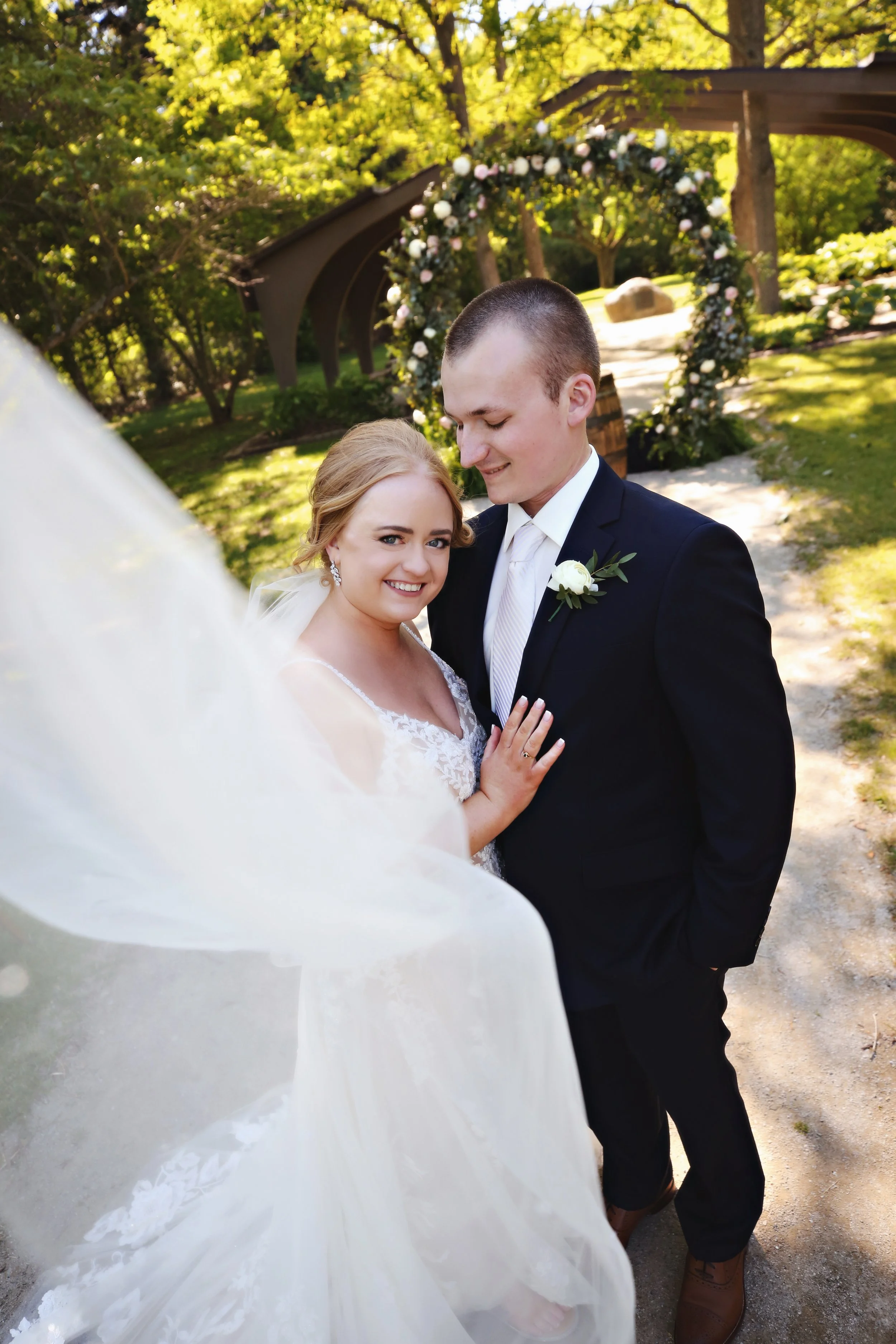 A smiling bride and groom posing outdoors on their wedding day, with a floral arch and lush greenery in the background.