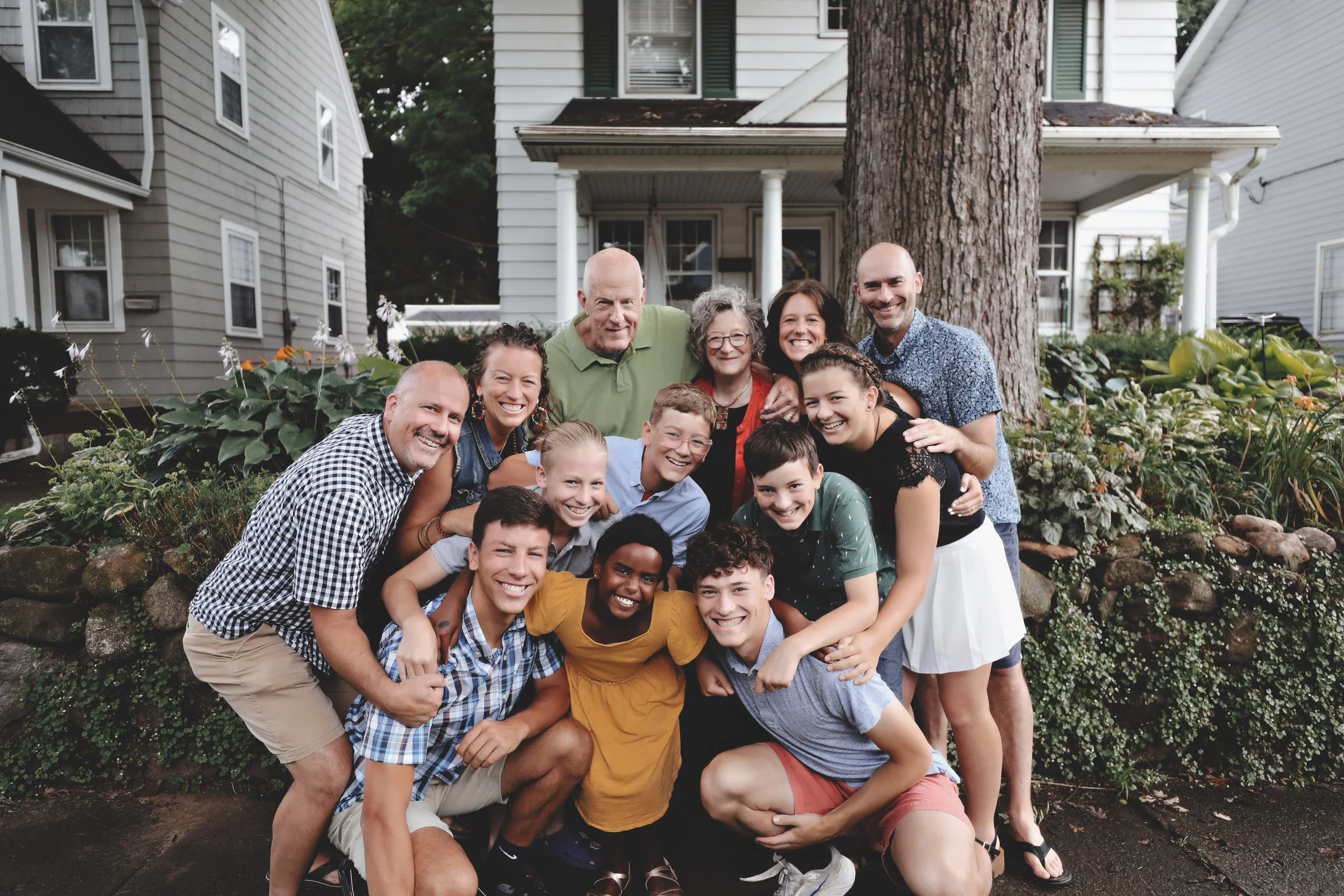 A large, diverse family group posing outdoors in front of a white house with a big tree and garden, smiling and hugging.