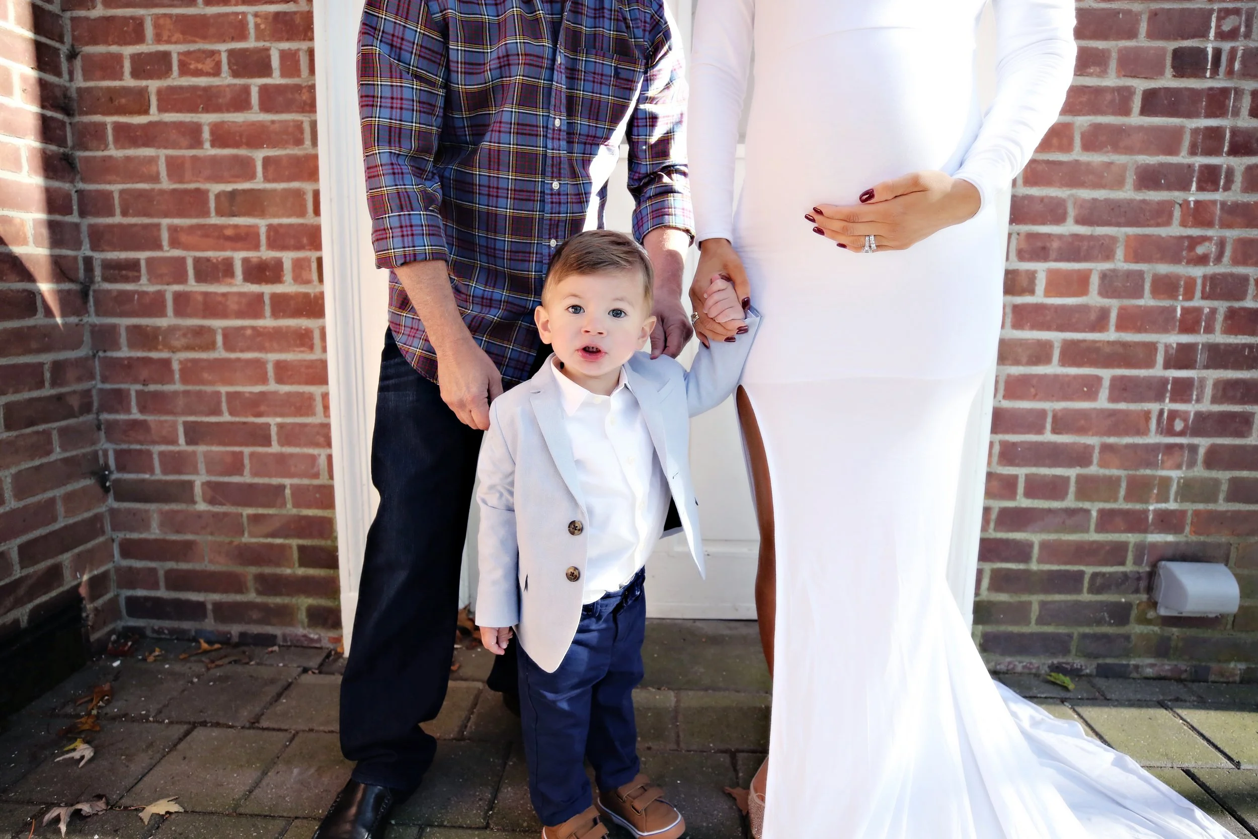 A young boy dressed in a white blazer, navy pants, and brown shoes, holding hands with a woman in a long white dress and a man in a plaid shirt, standing outside against a brick wall.