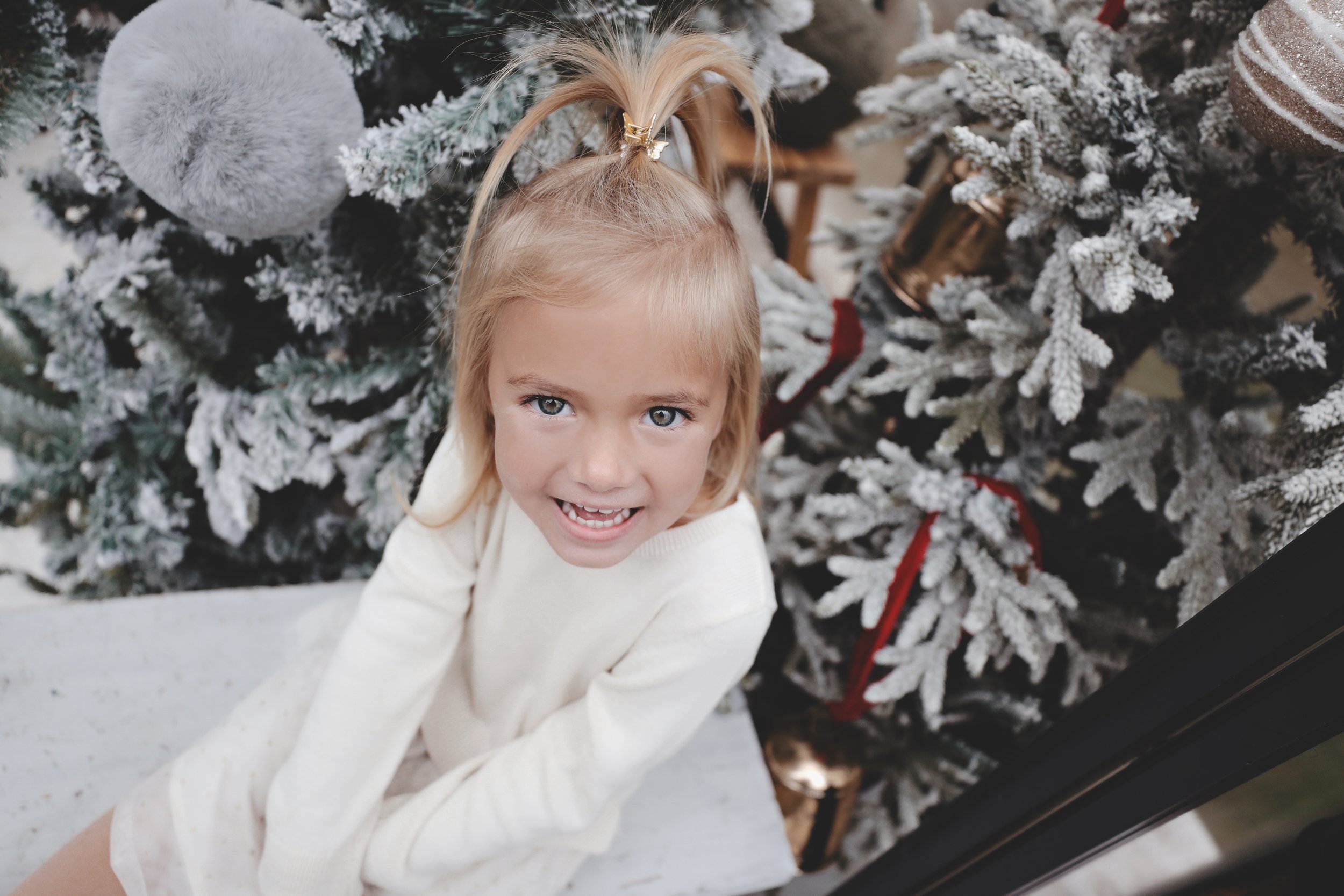 A young girl with blonde hair in a ponytail, smiling and looking up at the camera, standing in front of a decorated Christmas tree with snow-like flocking, ornaments, and ribbons.