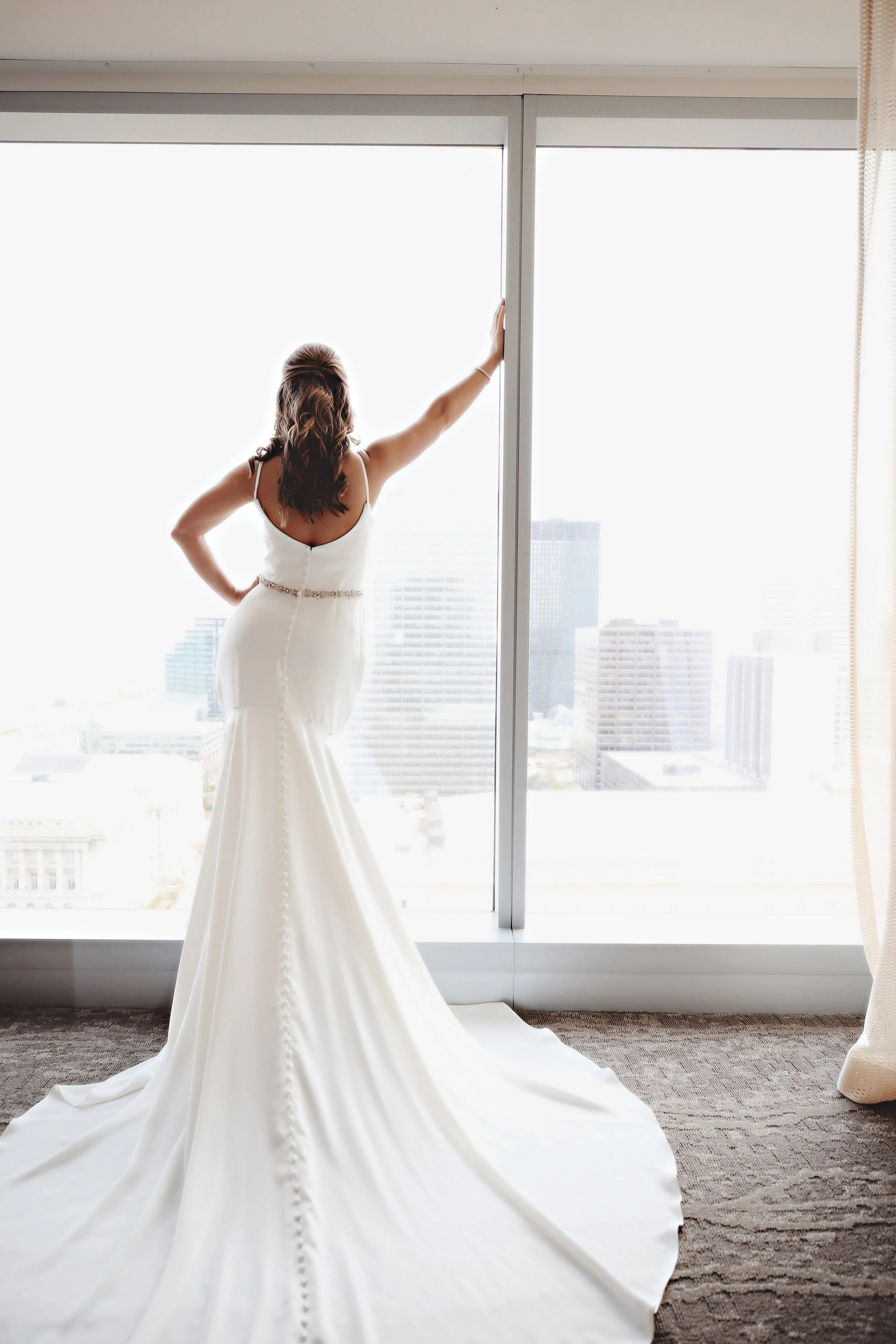 A woman in a white wedding dress stands inside a high-rise building, looking out through a large window at a cityscape with tall buildings, as she touches the window frame with one hand.