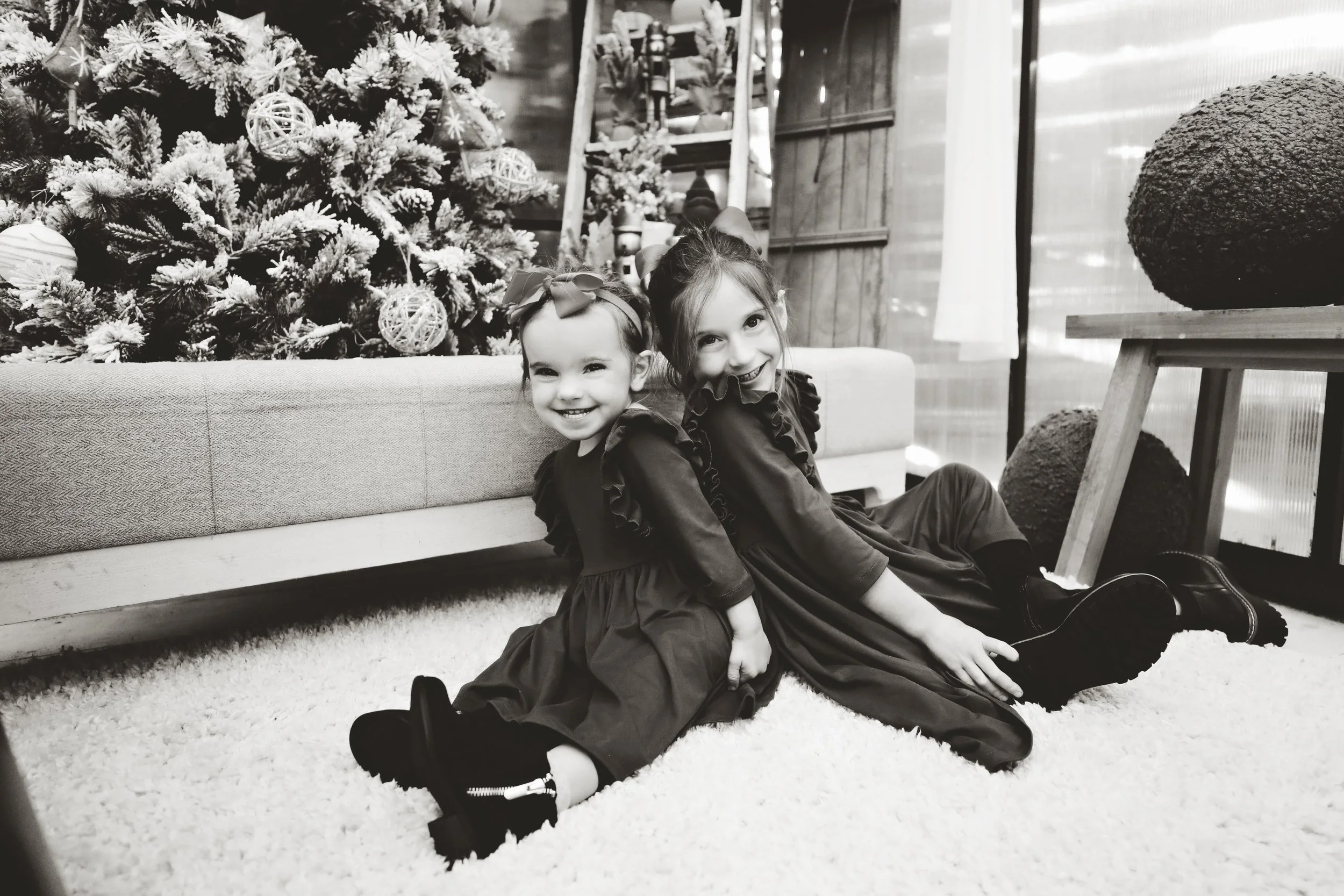Two young girls sitting back-to-back on a carpeted floor in front of a decorated Christmas tree, smiling at the camera. The girls are wearing dark dresses with ruffled sleeves and have bows in their hair, with one girl's hair tied up in a bun and the