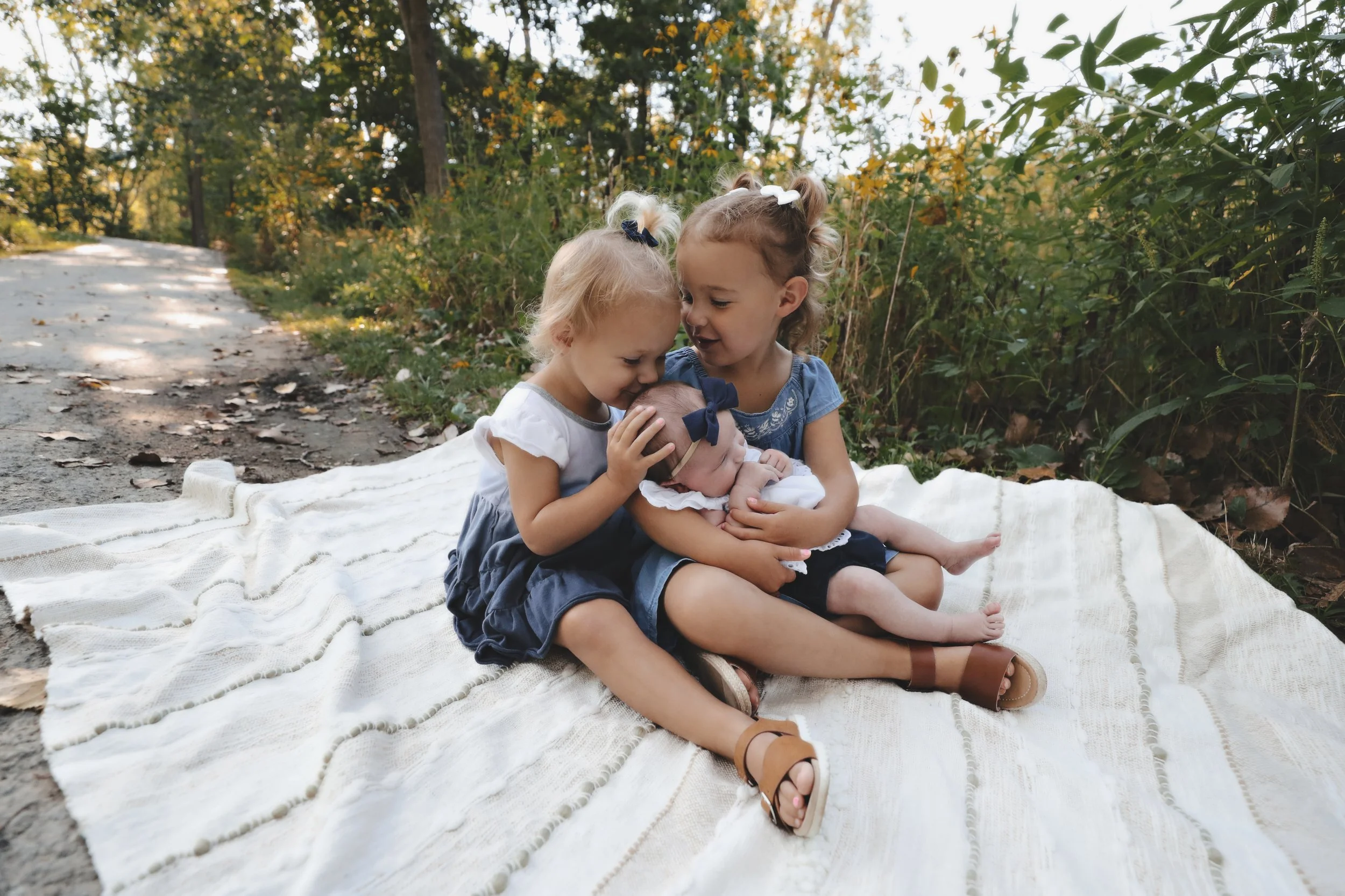 Three young girls sitting on a white blanket on the ground in a wooded outdoor area, with two older girls holding a baby girl, smiling and touching her forehead.