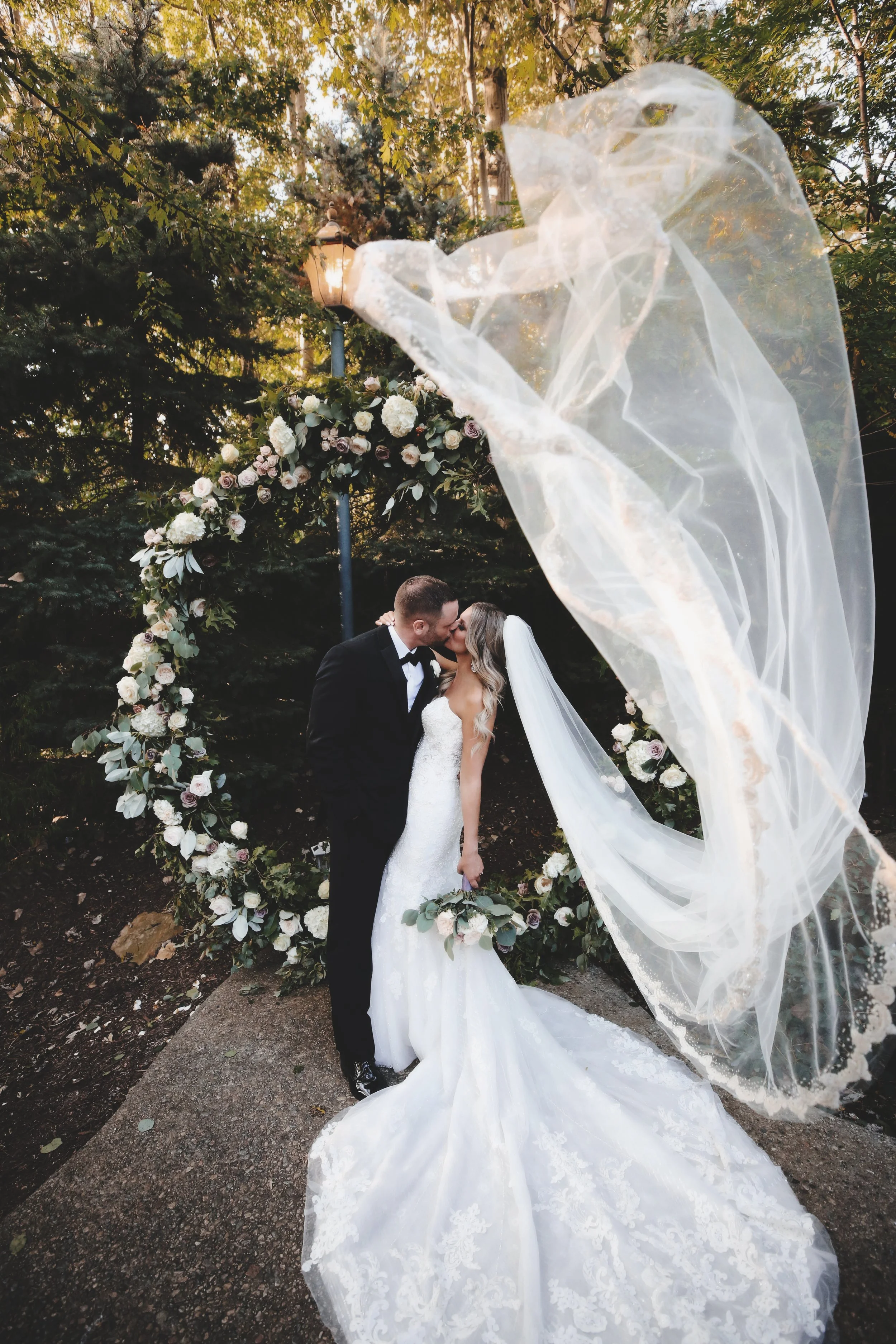 A bride and groom kissing under a floral wedding arch outdoors with trees and a lamp post in the background.