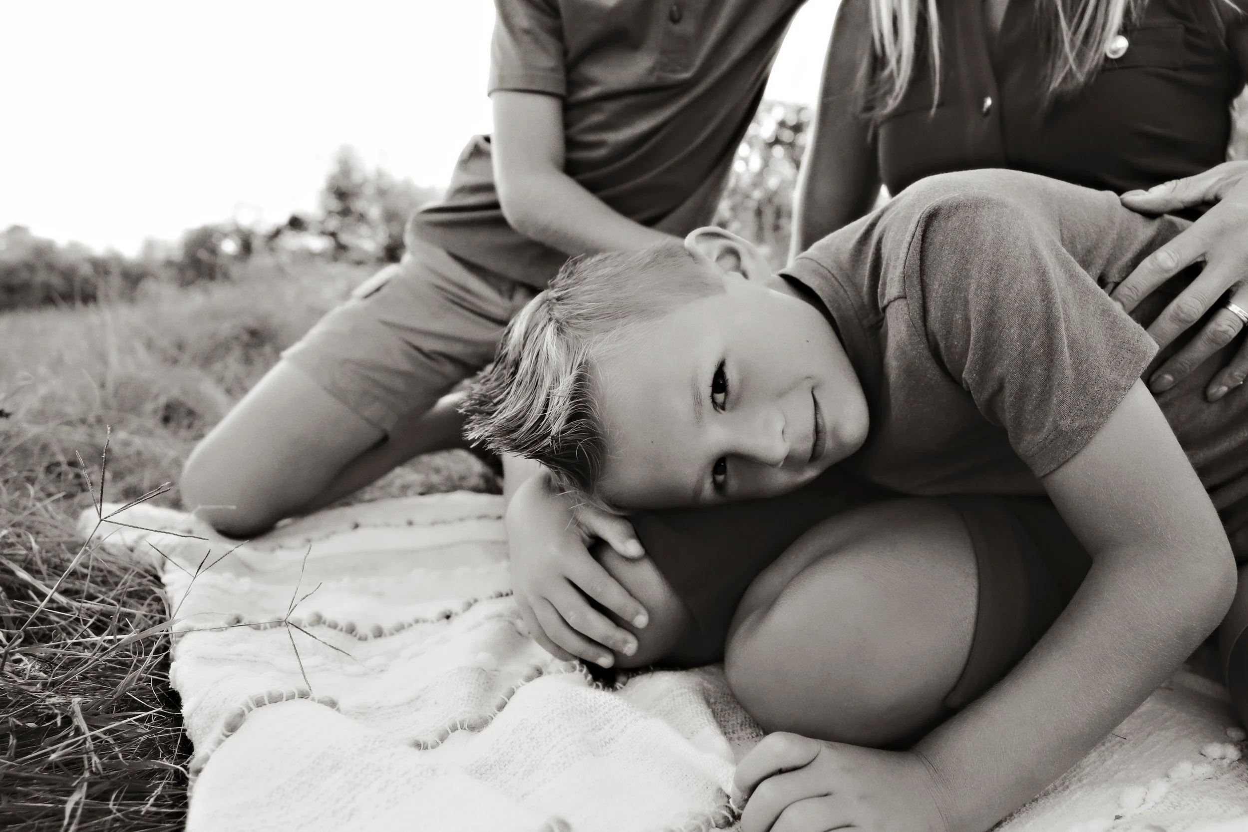 A young boy lying on the grass with a smiling face, resting his head on an adult's lap, while an adult sits nearby on a blanket outdoors.