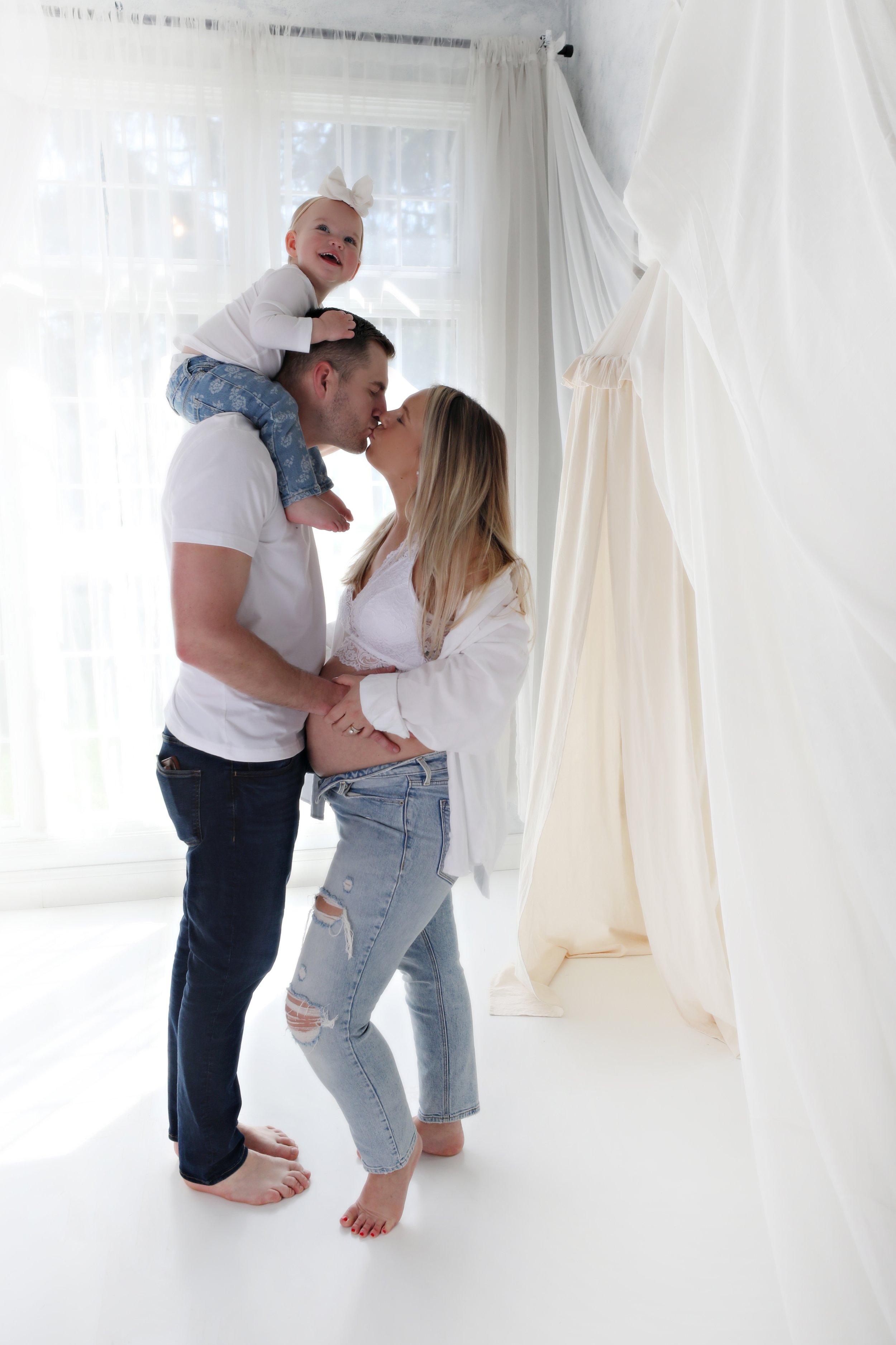 A family of three, a man, a woman, and a young girl, sharing a kiss indoors near a large window with white curtains. The girl is sitting on the man's shoulders, smiling and looking happy.