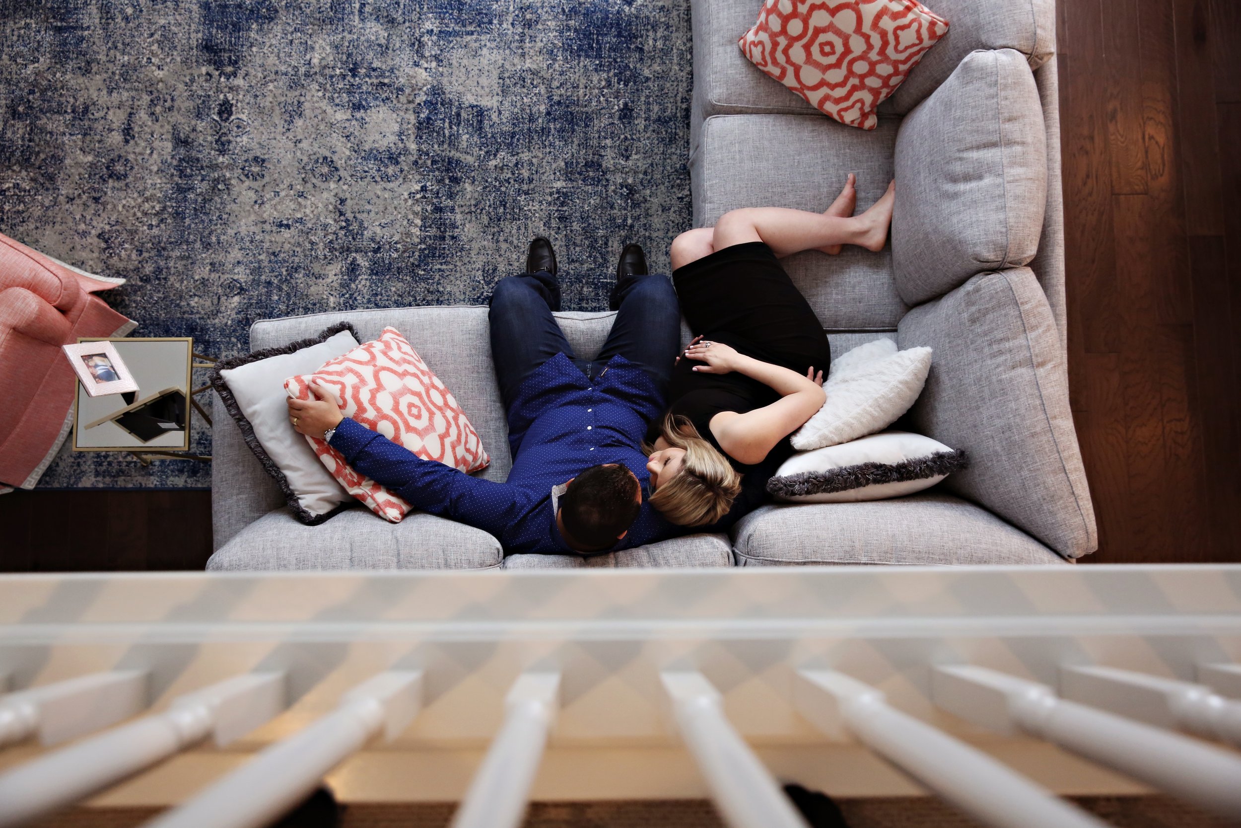 Overhead view of a couple lying on a gray sectional sofa, surrounded by pillows, in a living room with a blue patterned rug and wooden flooring.