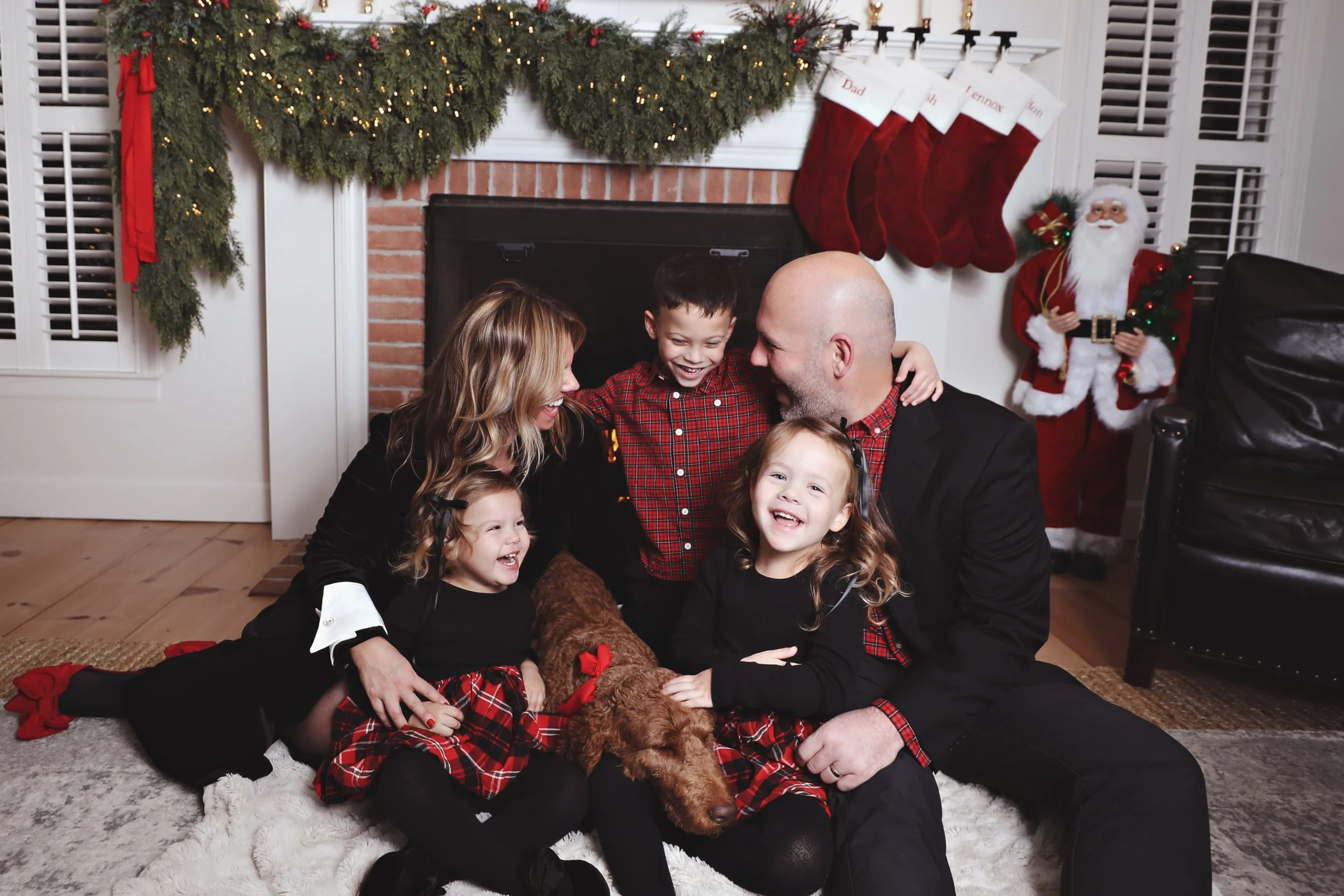 A family of five, including two young girls, a boy, and their parents, sitting on the floor in front of a fireplace decorated for Christmas with stockings hanging above and a Santa Claus decoration nearby. They are smiling and hugging a brown poodle.