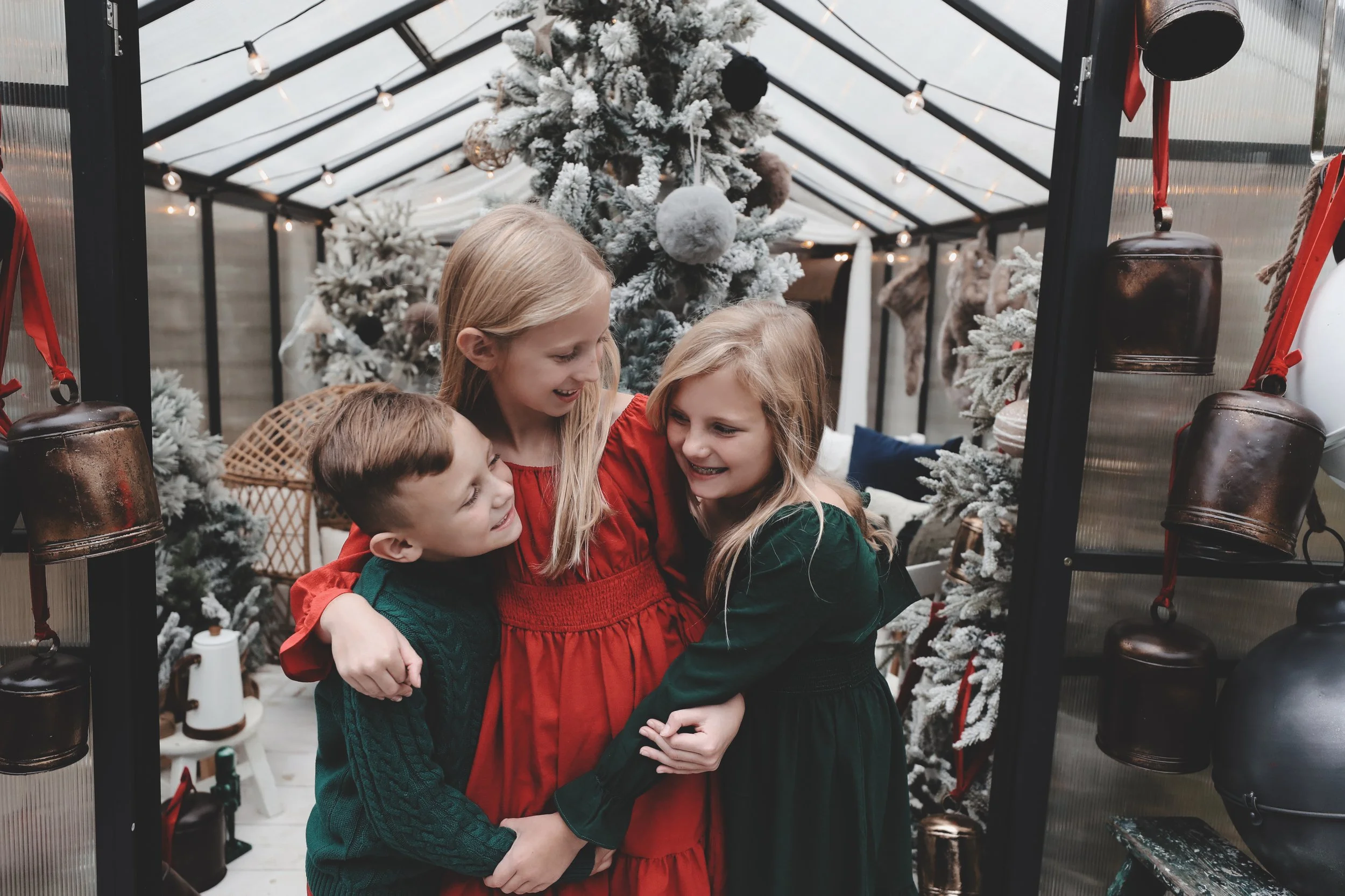 Four children, two girls and two boys, hugging and smiling in front of decorated Christmas trees inside a glass greenhouse.