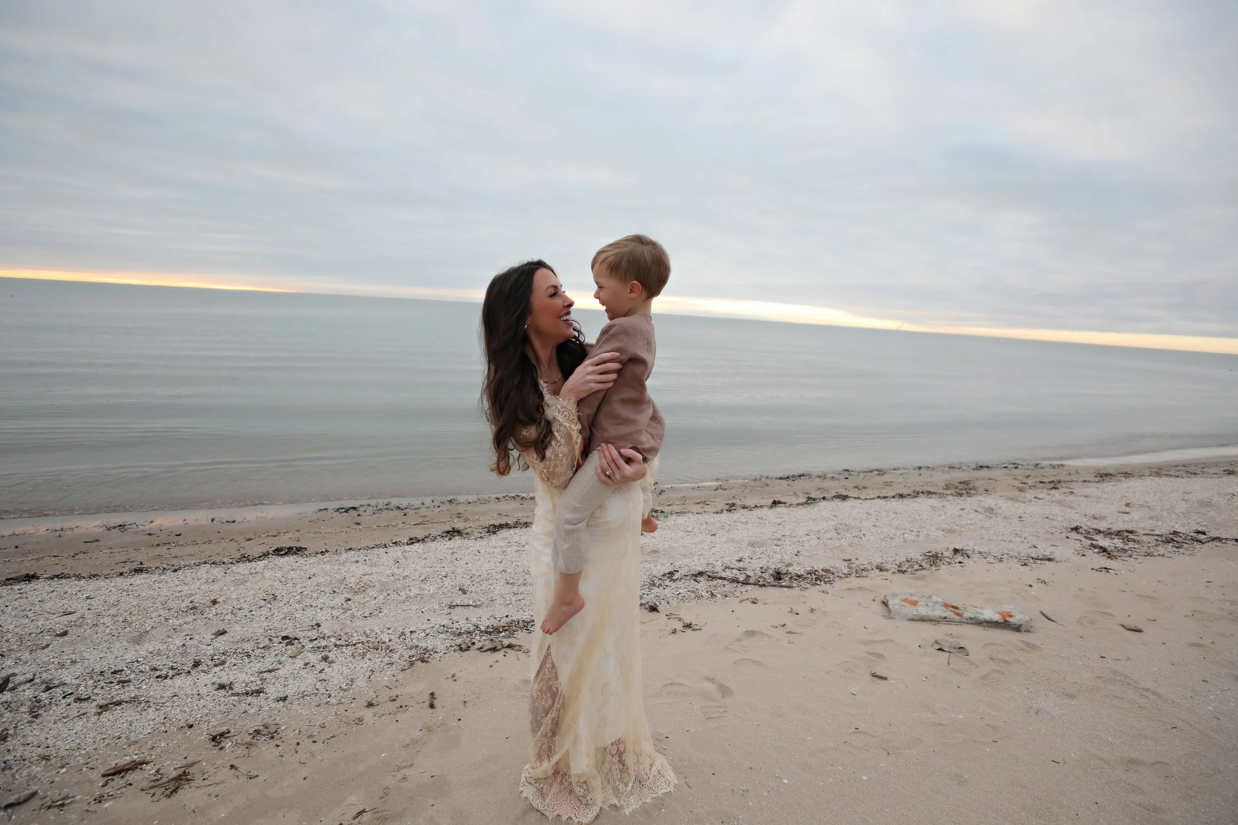 A woman in a cream-colored dress holding a boy in a brown jacket on a sandy beach at sunset, with the ocean and cloudy sky in the background.