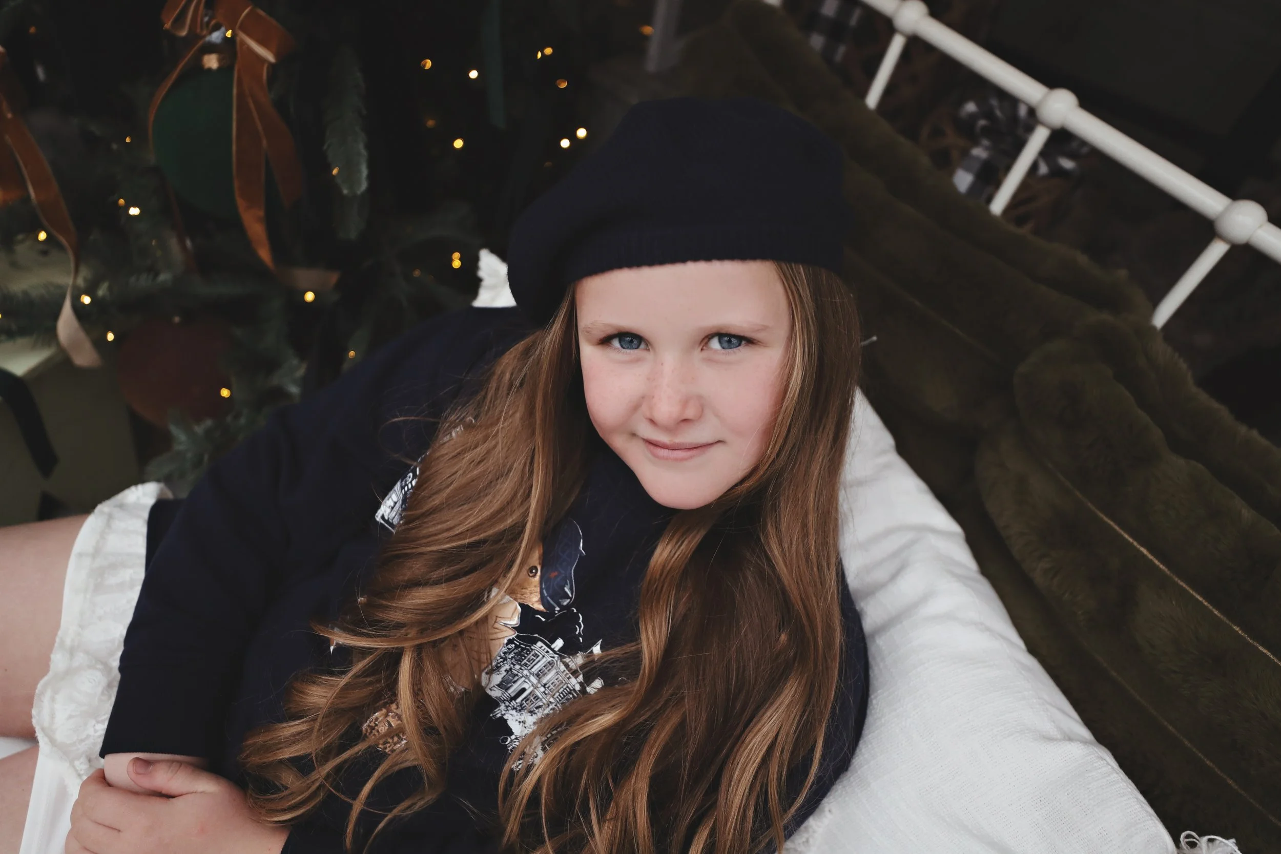 A young girl with long brown hair, wearing a black beret, lying on a white cushion, with a Christmas tree decorated with lights in the background.