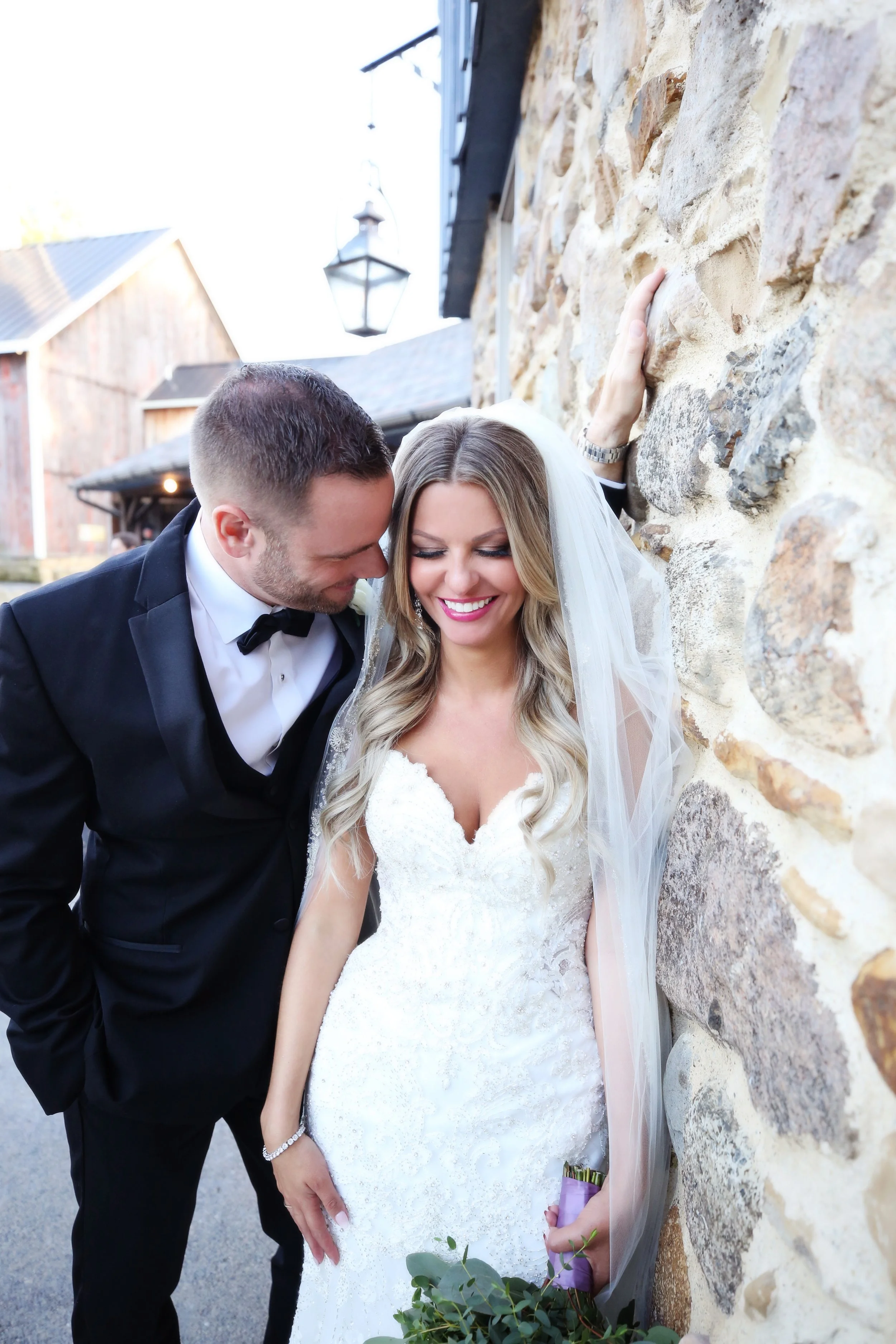 A newlywed couple in wedding attire smiling and leaning their heads together near a stone wall outside.
