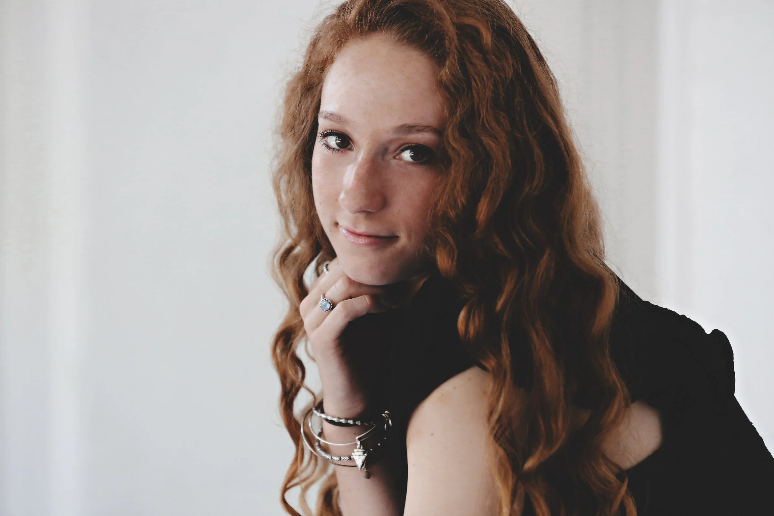 A young woman with long, curly red hair and fair skin, wearing a black sleeveless top, jewelry including rings, bracelets, and necklaces, resting her chin on her hand and smiling softly at the camera against a plain, light-colored background.