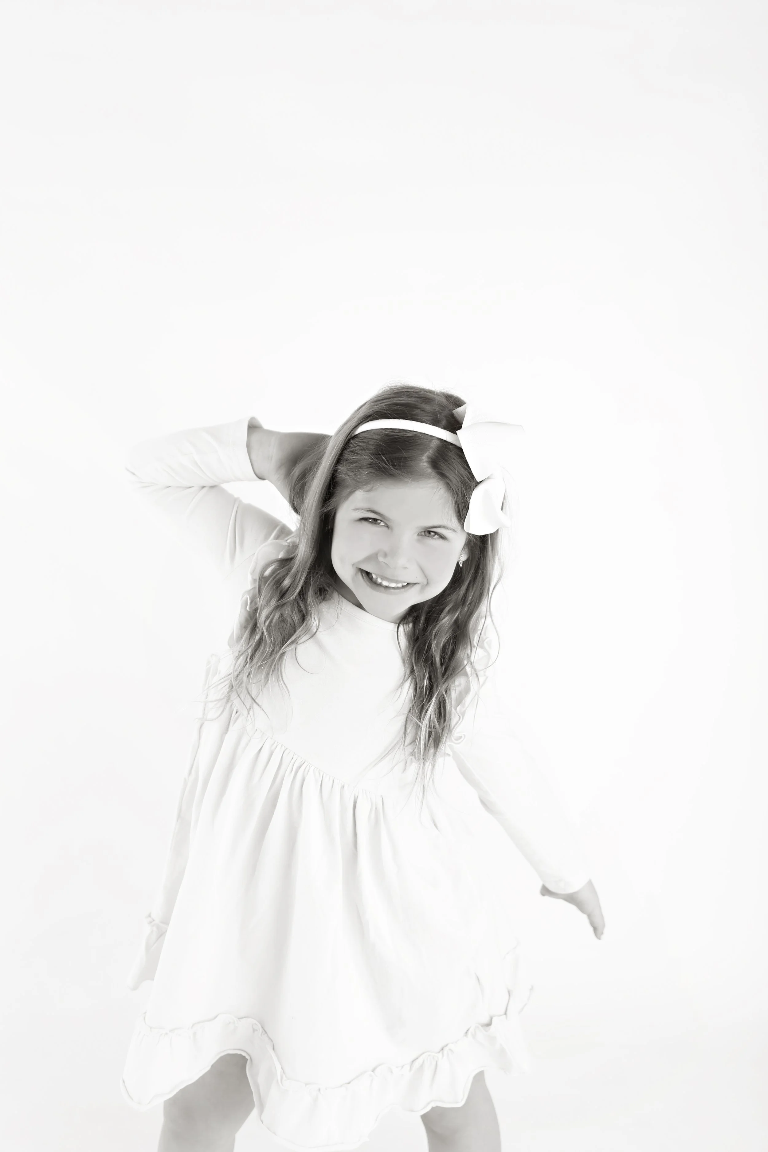 Black and white photo of a young girl with a happy expression, wearing a dress and a large bow in her hair, standing against a plain background.