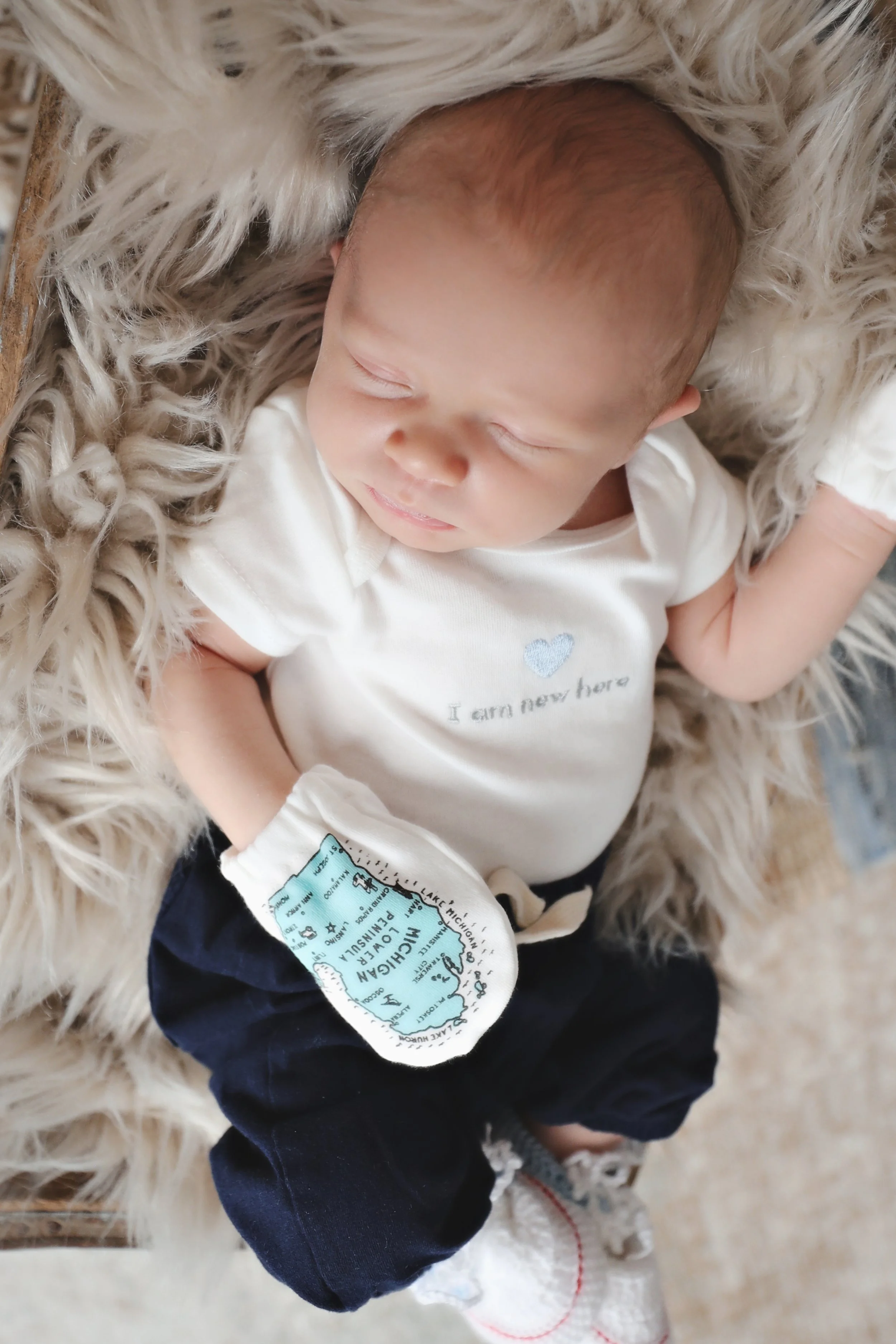 Sweet baby sleeping on a fluffy furry blanket, wearing a white shirt with a small heart and the words 'I am new here', navy pants, and a white glove with a Michigan map and hospital information.