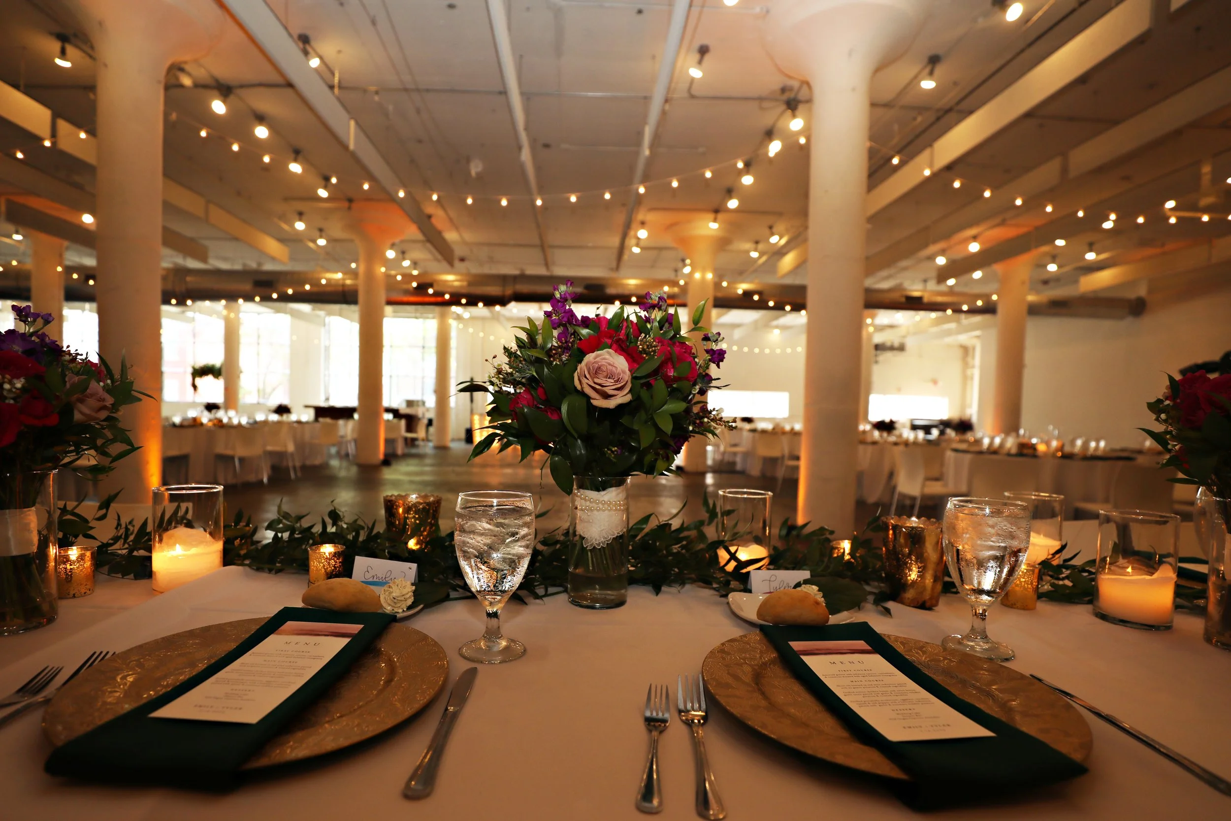 Elegant banquet table set with gold chargers, black napkins, wine glasses, and a floral centerpiece, in a spacious, warmly lit event hall with string lights and tall white columns.
