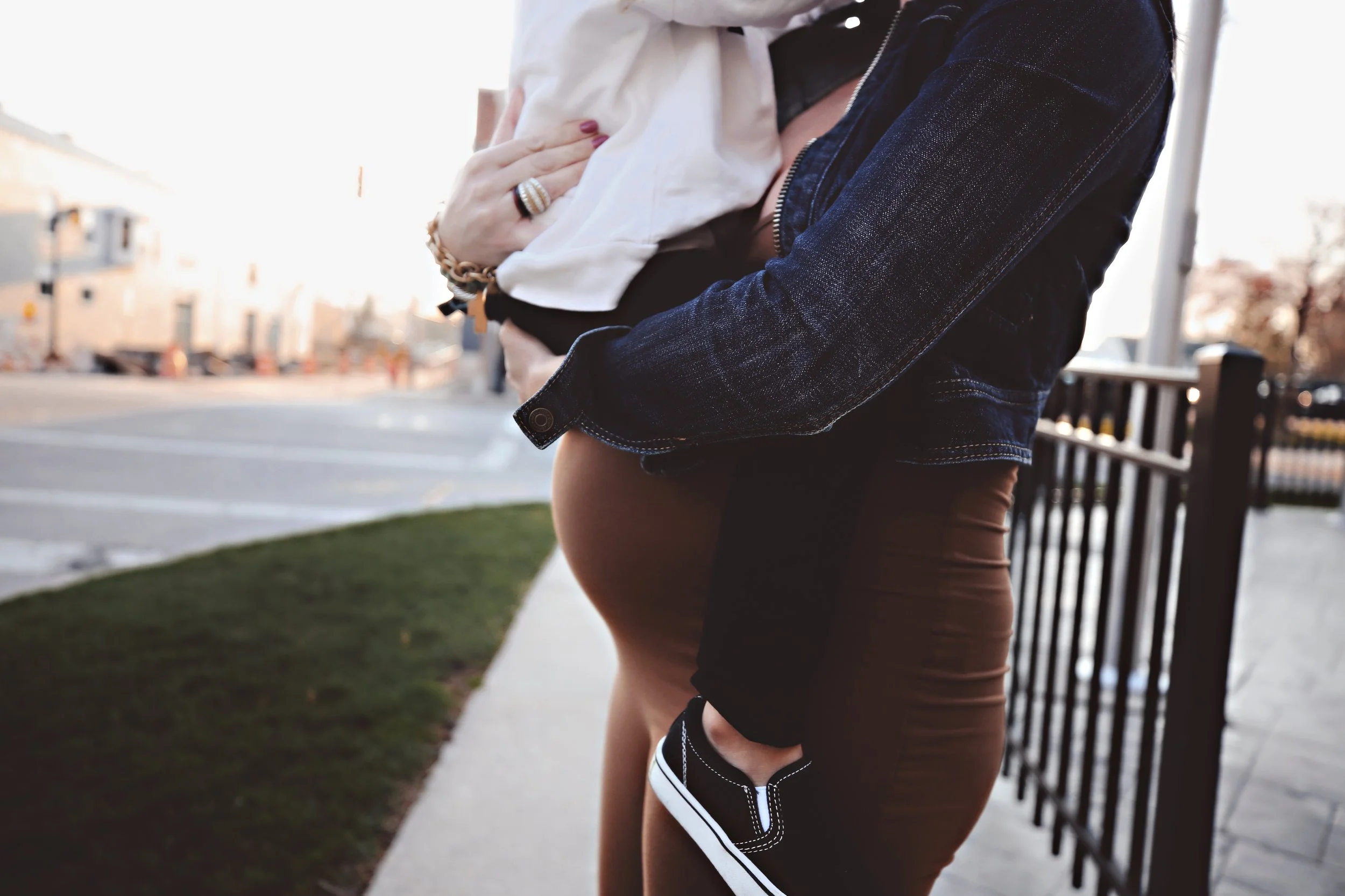A pregnant woman standing outdoors, holding a small child, with her hand on her belly, near a black metal fence and a sidewalk, in a neighborhood setting.