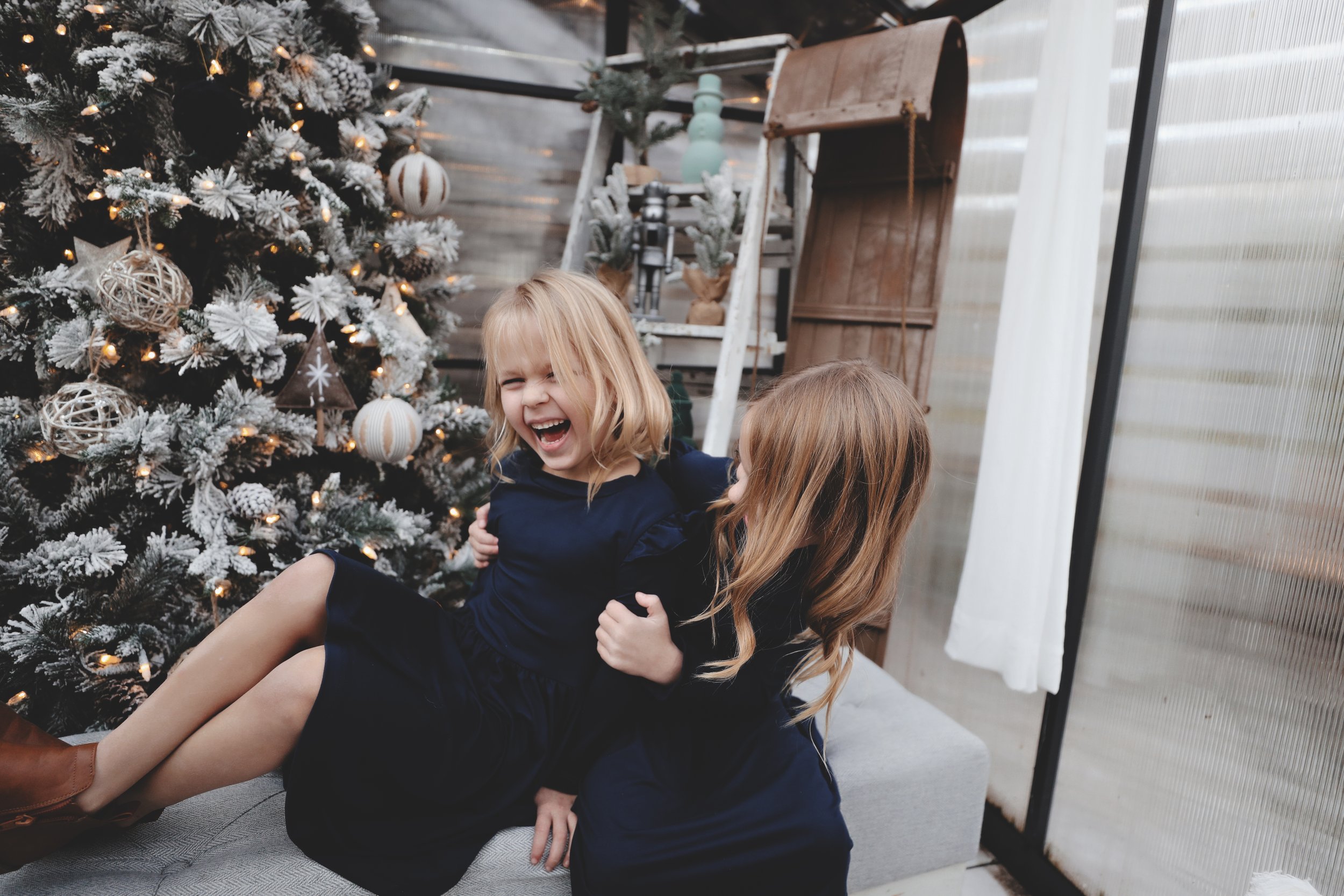 Two young girls in navy dresses smiling and laughing near a decorated Christmas tree in a cozy indoor setting.