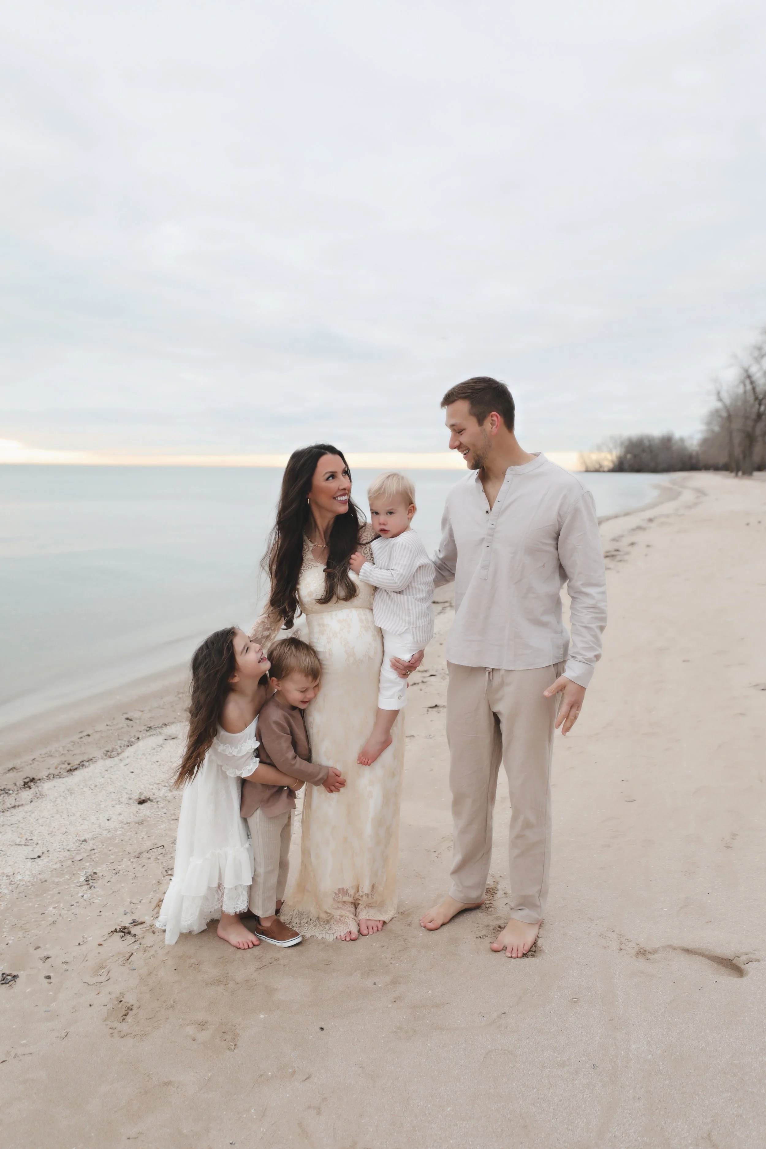 A family of five stands on a beach, smiling and interacting, with the ocean and cloudy sky in the background.