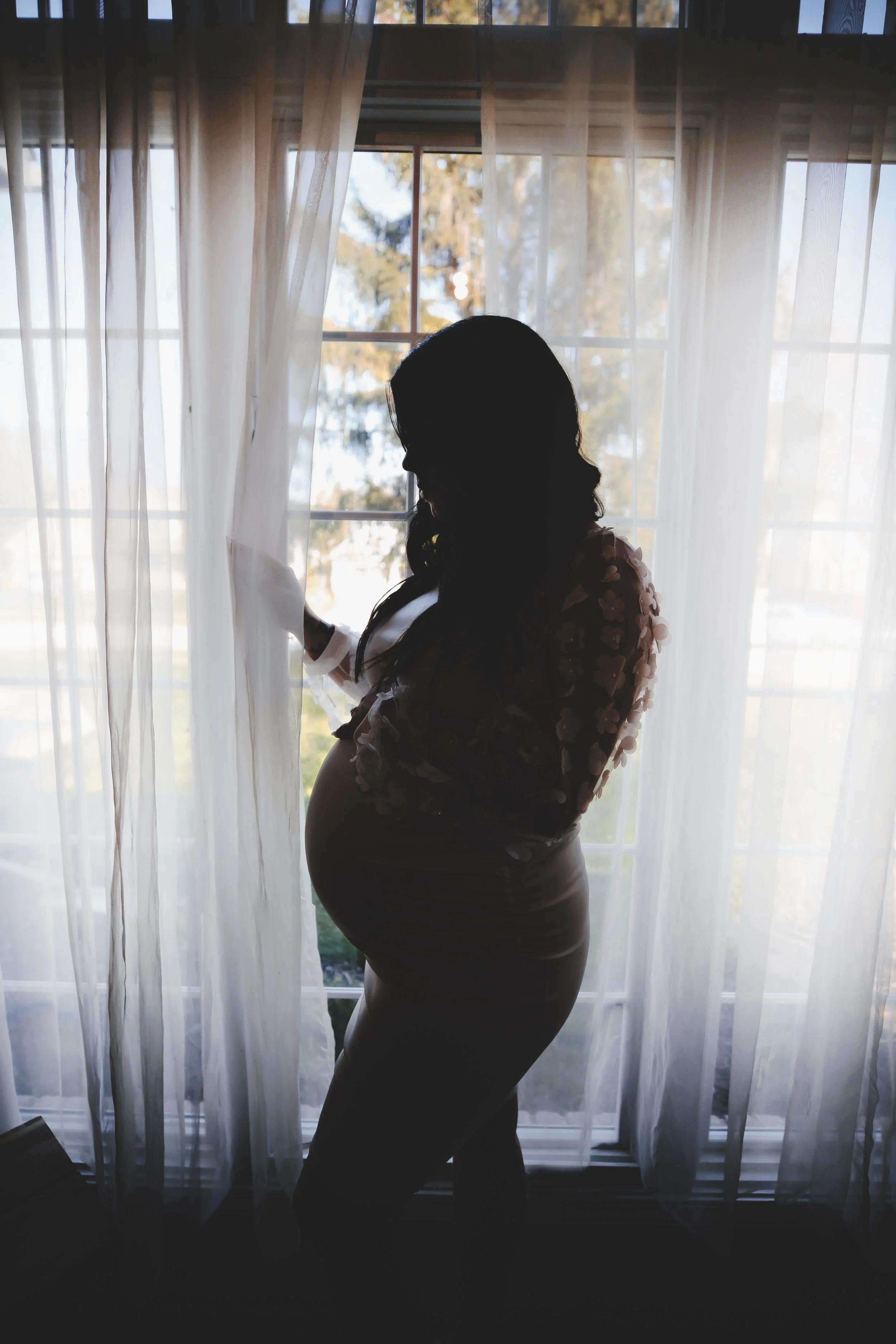 Silhouette of a pregnant woman standing by a window with sheer curtains, holding the curtains, backlit by sunlight.
