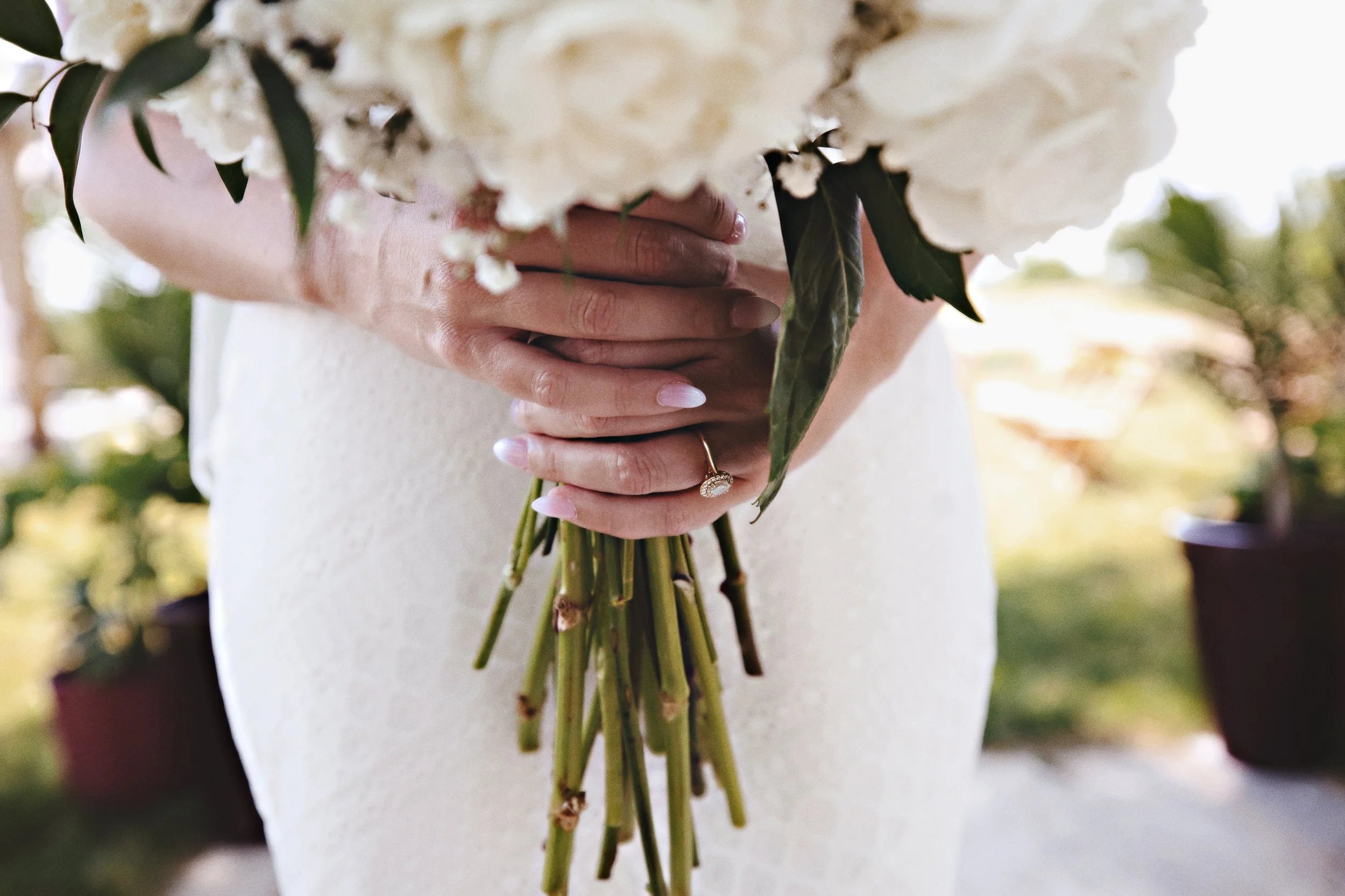 Person holding a bouquet of white flowers with green leaves, wearing a white dress and a ring on their finger.