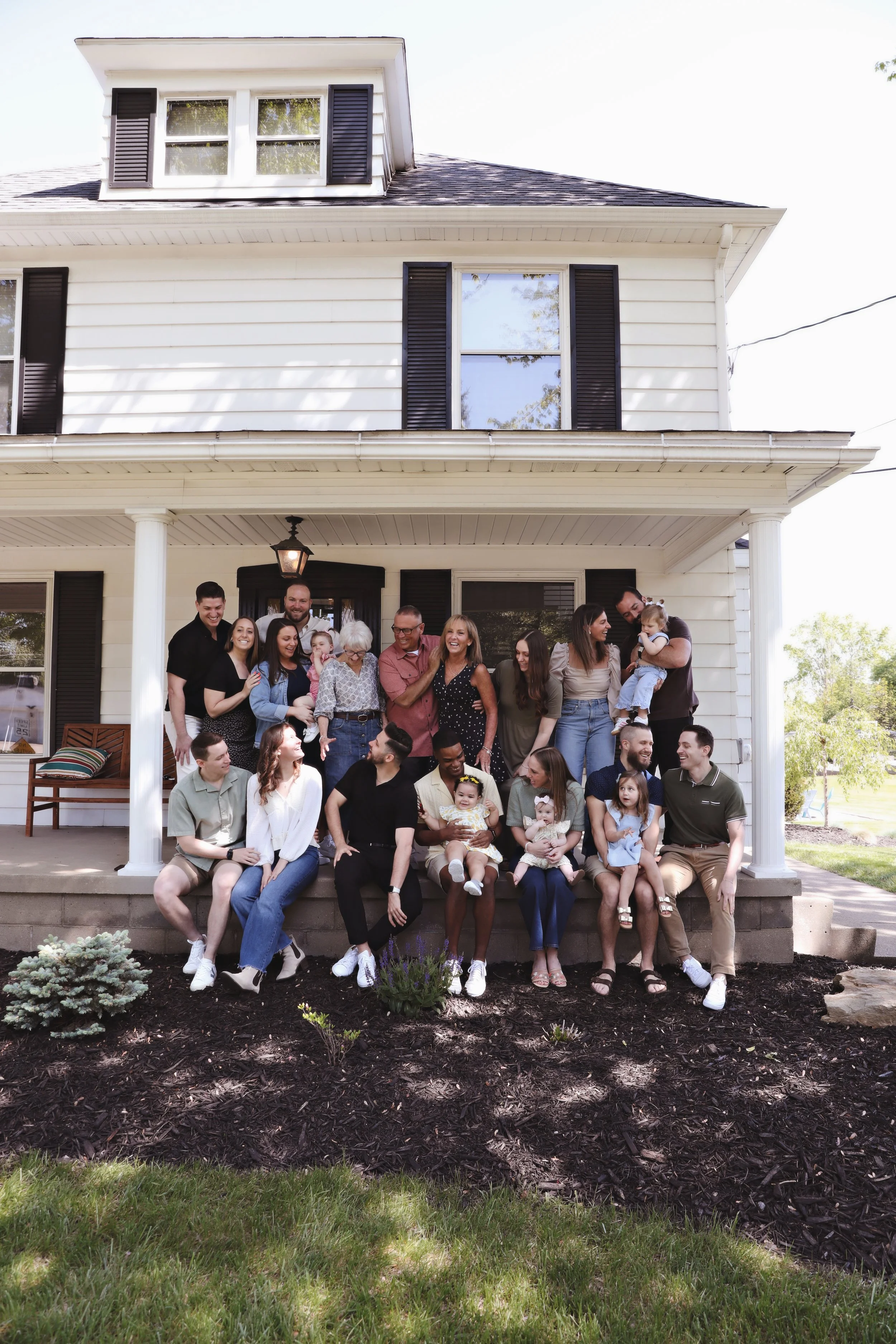 Large family gathering on the front porch of a white, two-story house with black shutters, with many adults and children sitting, standing, and interacting.