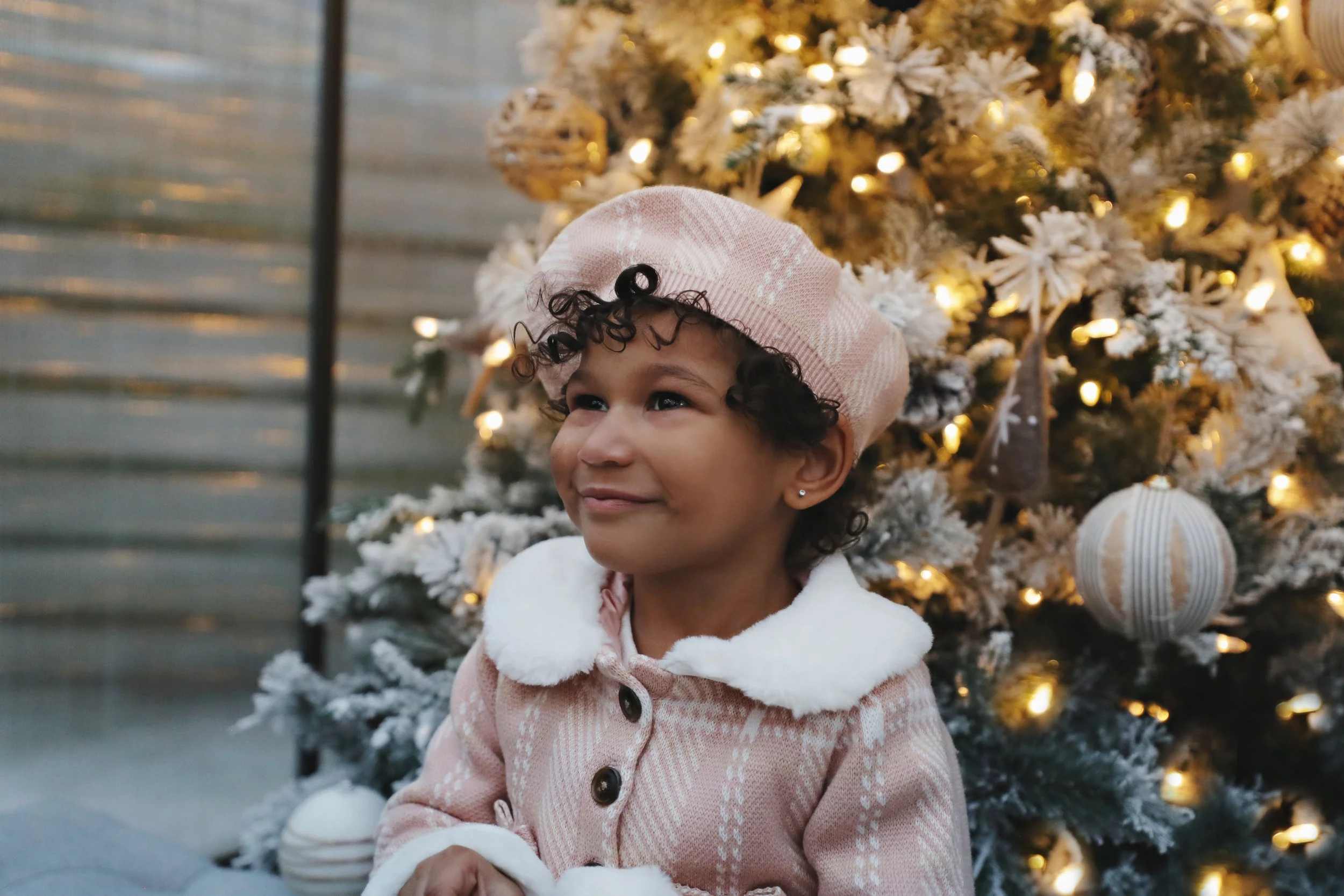 A young girl with curly hair wearing a pink beret, a pink coat with white fur trim, standing in front of a decorated Christmas tree with white ornaments and lights, smiling gently.