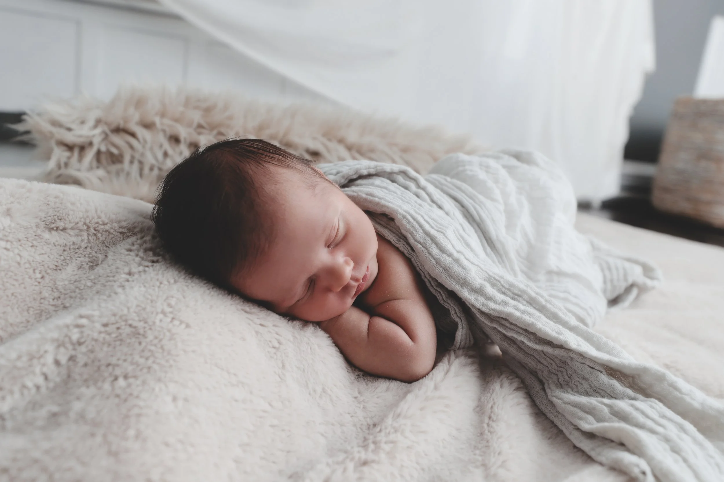 A sleeping newborn baby lying on a cream-colored, soft, furry blanket, covered partially with a white textured blanket, with a fluffy pillow in the background.