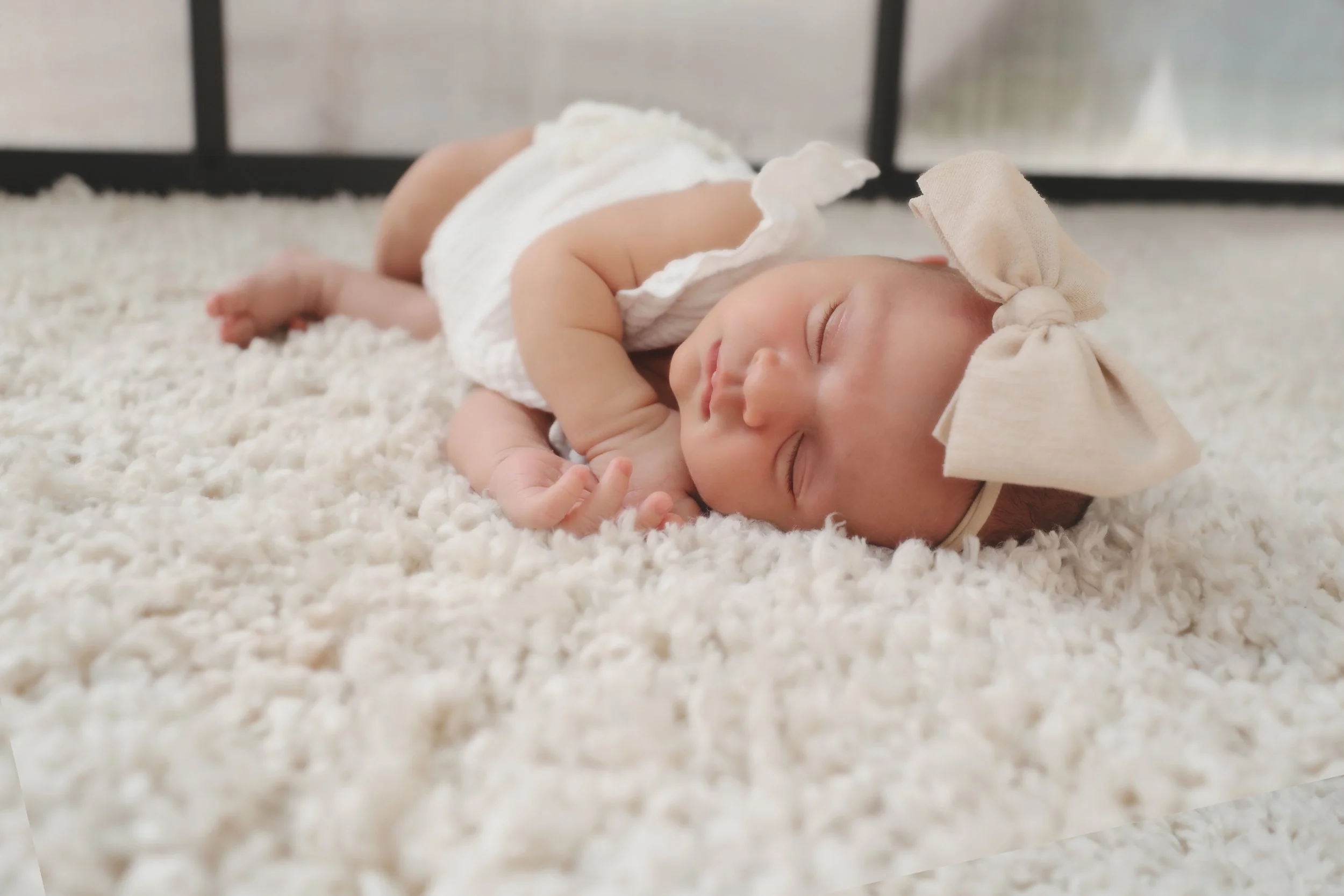 A sleeping baby girl with a large beige bow headband, lying on a soft white textured blanket.